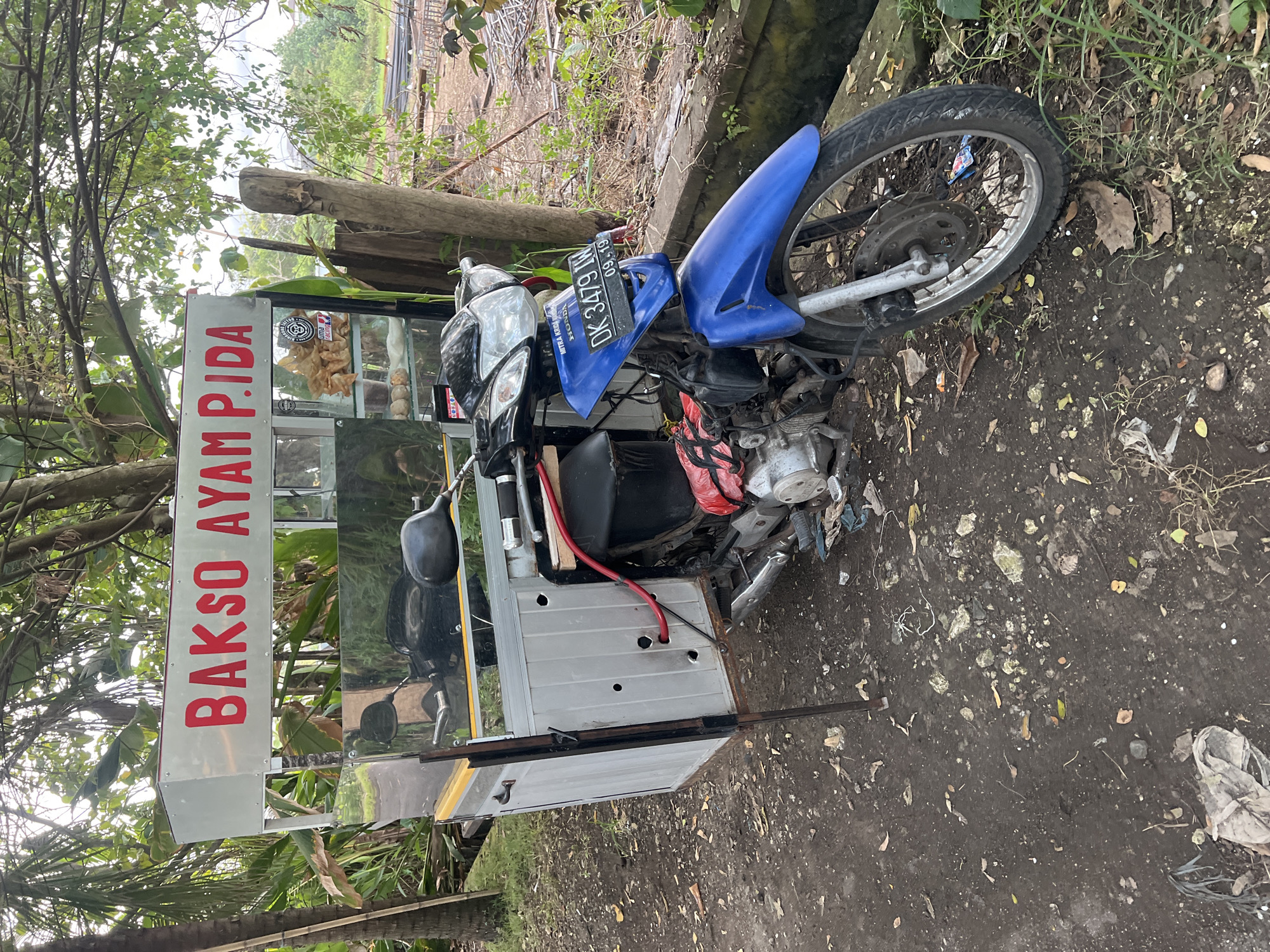 A food cart attached to a motorcycle sits on a dirt shoulder, possibly the side of a road. The cart features a sign that reads "Bakso Ayam P.IDA," suggesting it sells chicken meatball soup. Displayed inside the cart are food items, possibly related to the dish being sold. The motorcycle has a blue front fender and a visible license plate with the identification "DK 3479 IN" and an expiry date of 09.19. The surroundings consist of vegetation, potentially trees and shrubs, indicating an outdoor environment near the town of Mengwi, Indonesia.