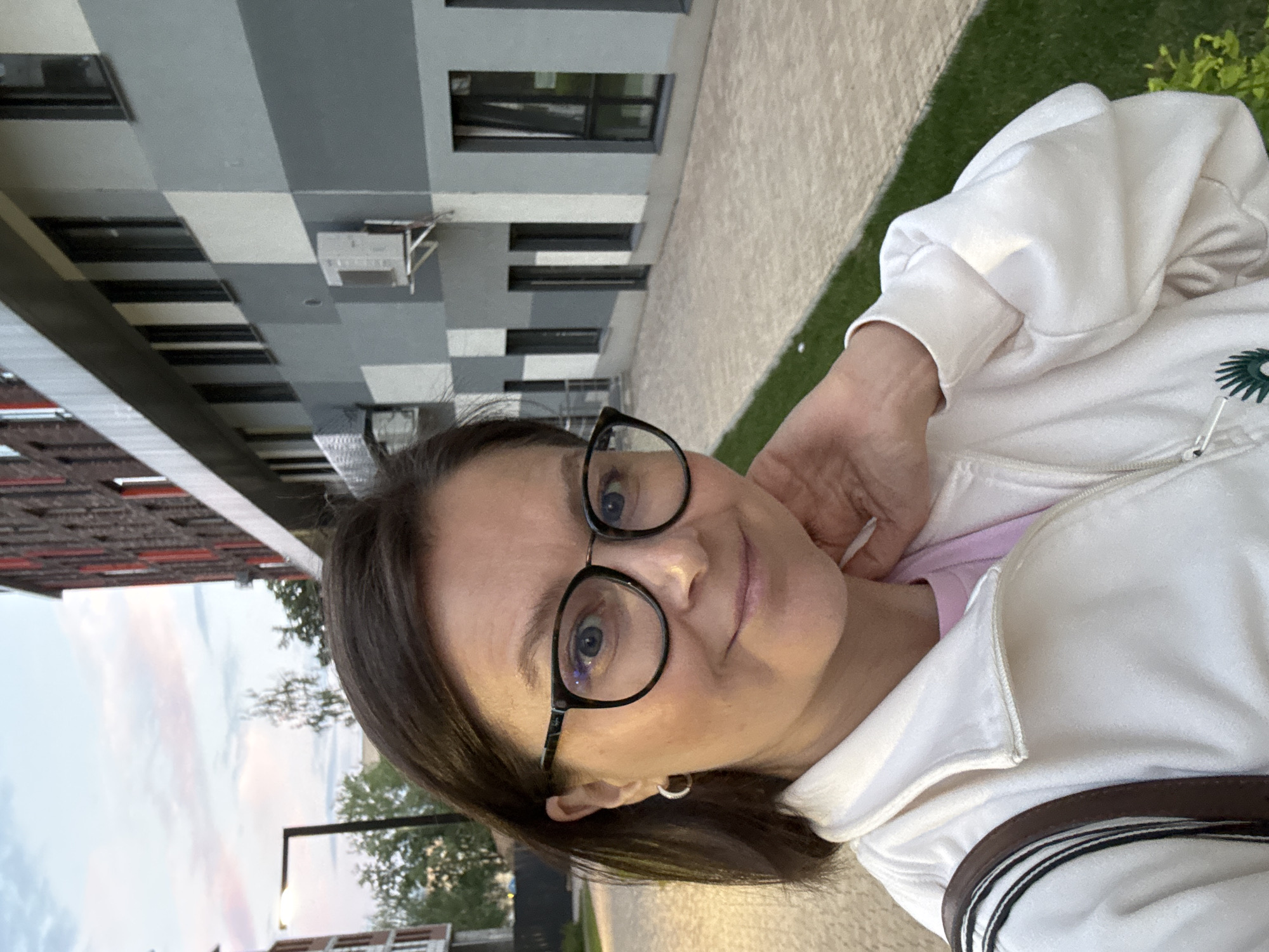 A woman with brown hair and glasses is taking a selfie outdoors. She is wearing a white zip-up jacket over a pink shirt, and a brown and black strap is visible over her shoulder. The background shows a modern building with a gray and white facade, with windows and air conditioning units visible. A paved walkway and a grassy area are in the foreground. The sky is overcast with hints of pink, suggesting it is either sunrise or sunset. The architectural style of the building and the overall appearance of the surroundings are consistent with contemporary urban development found in areas like Izhevsk, Russia.