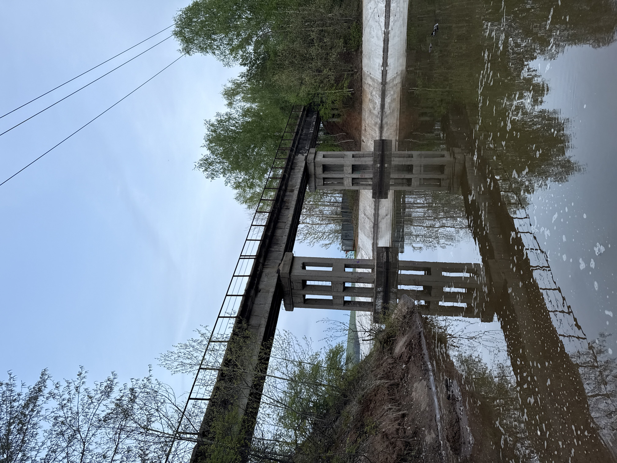 The image shows a concrete structure with a ladder on its side spanning a body of water. The structure appears to be a support or bridge. Its reflection is visible in the water. Trees and vegetation surround the structure on both sides. Power lines are visible in the upper left corner of the frame. The sky is overcast. The location is unidentified.