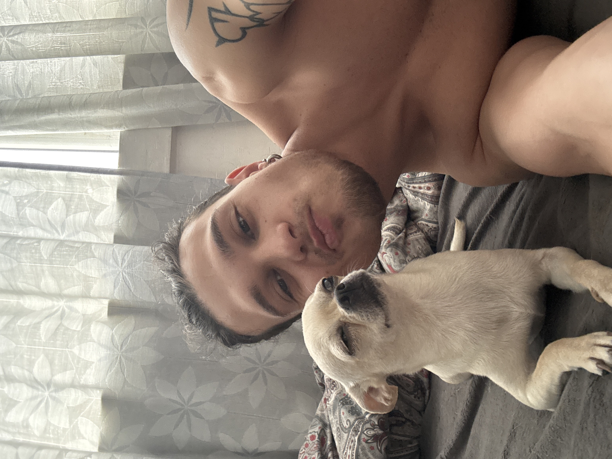 A man lies on a bed with a small, light-colored dog. The man has short, dark hair and stubble on his face. He is shirtless, revealing a tattoo on his shoulder. The dog lies close to the man's face. A window with sheer blinds is visible in the background. The image does not provide enough information to determine the location in Sofia, Bulgaria, or any specific actions or interactions.