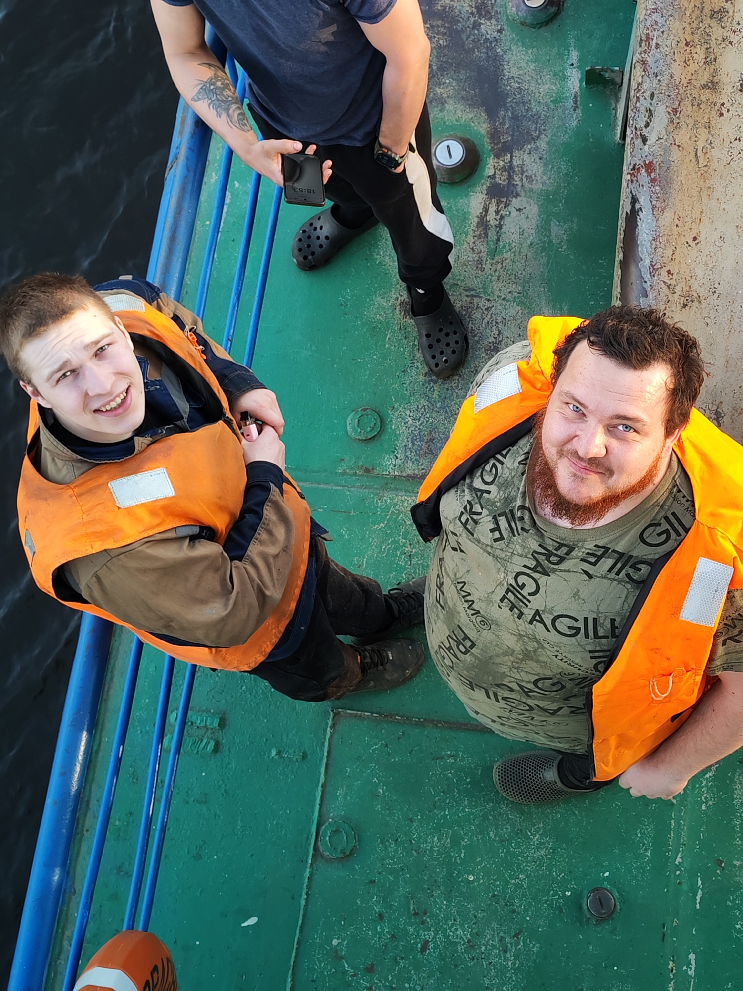 The image shows three men standing on a surface painted green, likely a deck of a boat or ship. The location is possibly in or near Базарово, Russia, though this is not definitively proven by the image itself.
Two of the men are wearing orange life vests. The first man is younger, with short brown hair, and is smiling at the camera. He is also holding a small pink object in his hand. The second man is older, with a red beard, and is also looking at the camera. He is wearing a green t-shirt with the words "FRAGILE" and "AGILE" printed repeatedly on it.
The third man is partially visible in the upper part of the frame. He is wearing a dark blue shirt and black pants with white stripes on the side. He has a tattoo on his left arm and is holding a mobile phone with the screen showing "18:53".
The men are standing next to a railing painted blue. Beyond the railing is dark water. The surface they are standing on shows signs of wear and age. There are several metallic circular objects embedded in the green surface.