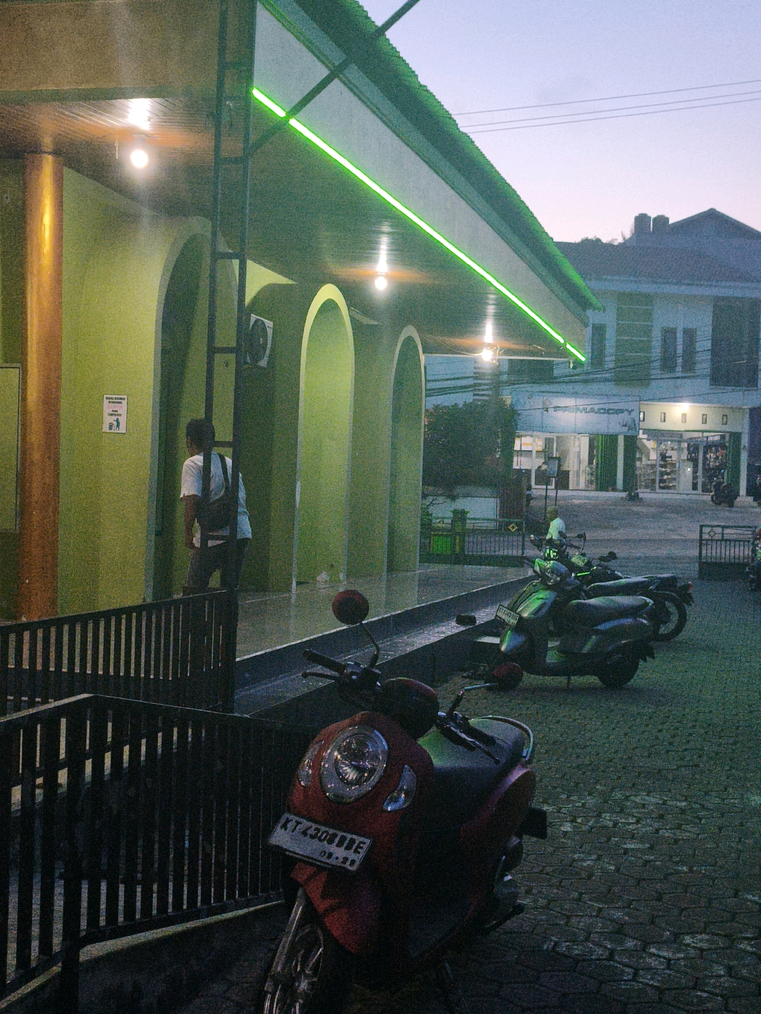 The image depicts an outdoor scene in front of a building with a green roof and archways, possibly a mosque or a public building, in Samarinda, Indonesia. The building's walls are painted a light green, and there is a bright green neon light strip running along the edge of the roof. Several light bulbs are visible on the underside of the roof. In the foreground, a person wearing a white t-shirt and dark pants, carrying a backpack, is walking away from the camera. A black metal fence is in the lower left of the image. Several motorcycles are parked on a cobblestone surface in the middle and right side of the frame. The sky is a pale twilight blue. In the background, a multi-story building with windows and a sign that reads "PRIM COPY" is visible, suggesting a commercial area. A few trees and power lines are also present in the background. The overall lighting suggests it is dusk or early evening.