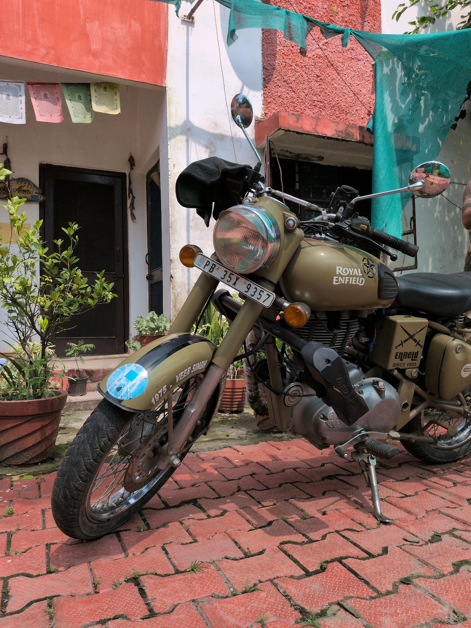 An olive green Royal Enfield motorcycle is parked on a red brick-patterned pavement in front of a two-story building. The building has a red stucco facade on the upper story and a white lower story. Prayer flags are hung from the overhang of the building. A tree in a large terracotta pot is to the left of the motorcycle, and other potted plants are visible. The motorcycle has a license plate that reads "PB 35X 9357." The motorcycle also has "Royal Enfield" written on its fuel tank and "VEER SINGH" and "1975" on the front fender. A green net is draped over the right side of the motorcycle and the building. The scene appears to be in Pathankot, India, based on the license plate format.