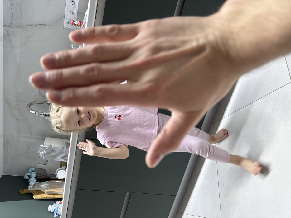 The scene, located in Gornyi (Dvurechenskoe S/P), Russia, depicts a domestic kitchen interior. A blurred, light-skinned adult's right hand occupies the immediate foreground, palm facing forward with fingers slightly spread. Behind the hand, a fair-skinned female child, estimated to be between 3 and 5 years old, smiles towards the camera. She has blonde hair styled in two pigtails and wears a light pink short-sleeved t-shirt with a red cherry graphic on the chest, along with matching light pink leggings. She stands barefoot on a light grey tiled floor, with her left foot partially lifted, suggesting motion. Her right hand is raised, palm facing forward, appearing to engage in a reciprocal gesture with the foreground hand. The background reveals dark green kitchen base cabinets topped by a light-colored countertop and a tiled backsplash. A chrome kitchen faucet is visible above a sink. Various objects are present on the countertop, including a wooden cutting board, glass bottles, a clear pitcher, and a white power outlet with a small decorative figure.