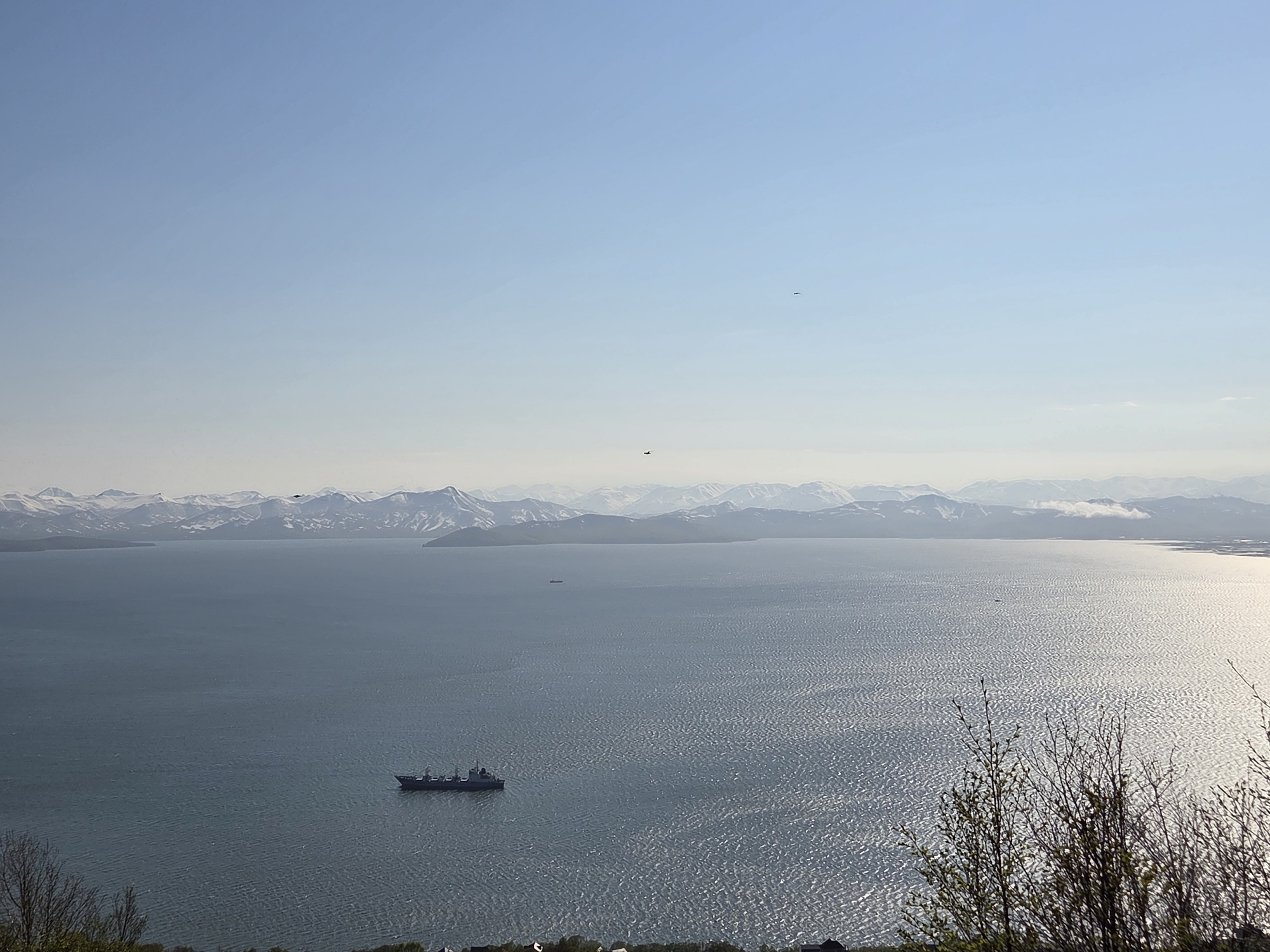 The image depicts a wide-angle view of a bay or body of water with a distant, snow-capped mountain range in the background. The water is calm with a rippled surface reflecting the sunlight. A dark-colored ship, appearing to be a cargo vessel or a smaller naval ship, is visible in the middle ground, to the left of the frame. The sky is clear and pale blue, with a few faint white clouds near the horizon. In the foreground, there are some bare tree branches and sparse greenery, suggesting it might be spring or early summer. The setting appears to be a scenic coastal area, likely overlooking the bay near the city of Krutoberegovy, Russia, known for its natural beauty and proximity to the sea. There are no visible people or discernible actions taking place in the scene.
