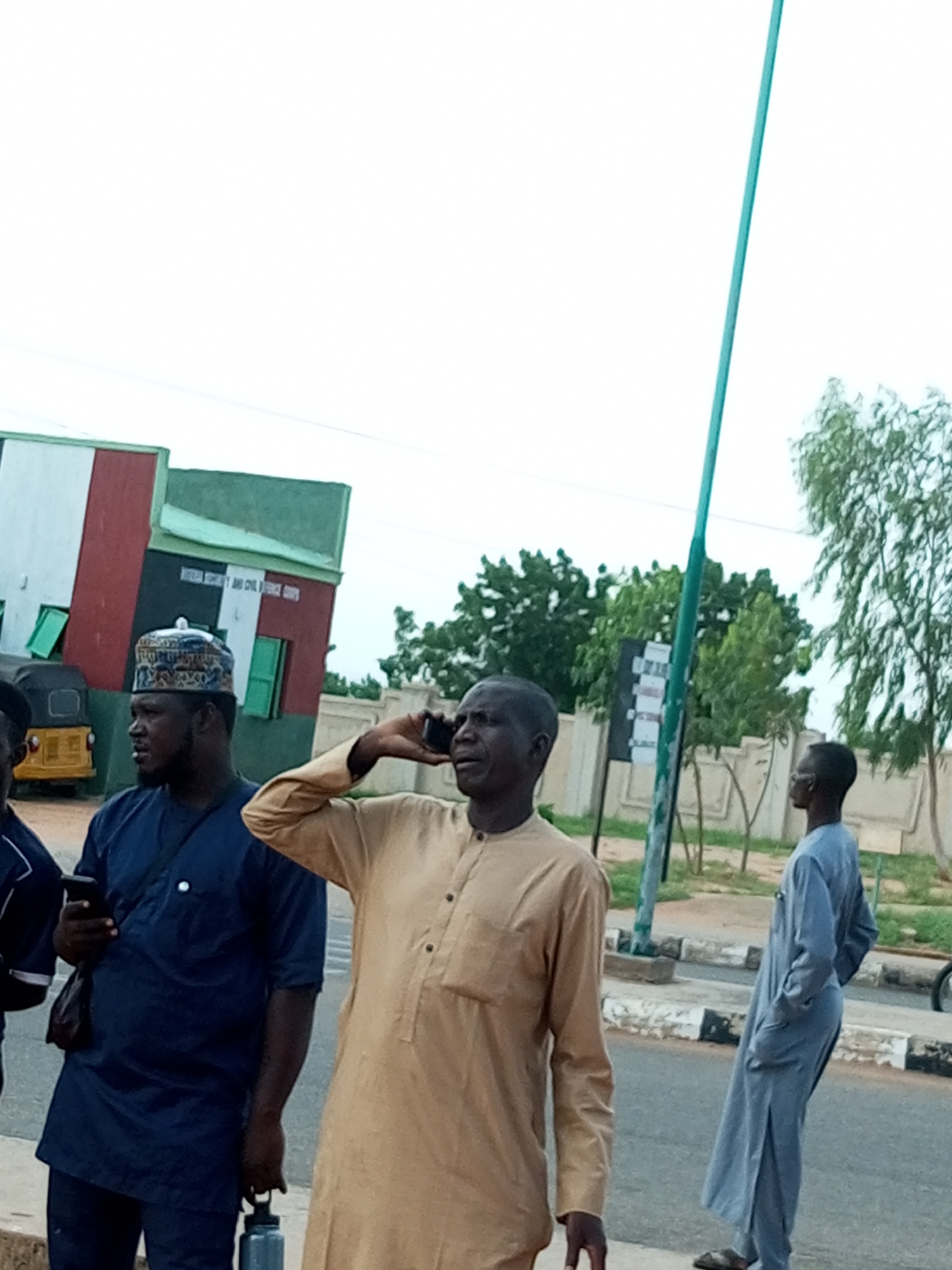 The image depicts a street scene in Gombe, Nigeria, with several individuals present. In the foreground, a man in a light brown robe is holding a mobile phone to his ear, engaged in a conversation. To his left, another man in a dark blue outfit, wearing a patterned cap, is looking to the side and holding a smartphone in his left hand. He is also carrying a dark bag over his shoulder and a grey water bottle in his right hand. Further to the right, a man in a light blue robe stands with his hands in his pockets, facing away from the camera.
In the background, a building with a distinctive facade is visible, featuring red, white, and black vertical stripes, with green accents. Text on the building appears to read "NATIONAL SECURITY AND CIVIL DEFENCE CORPS". A yellow auto-rickshaw is parked to the left of the building. The setting appears to be outdoors, with trees and a street with a curb visible. The sky is overcast, suggesting cloudy weather. The overall atmosphere seems to be one of casual interaction or waiting. The time of day is not explicitly indicated but the lighting suggests it is daytime.