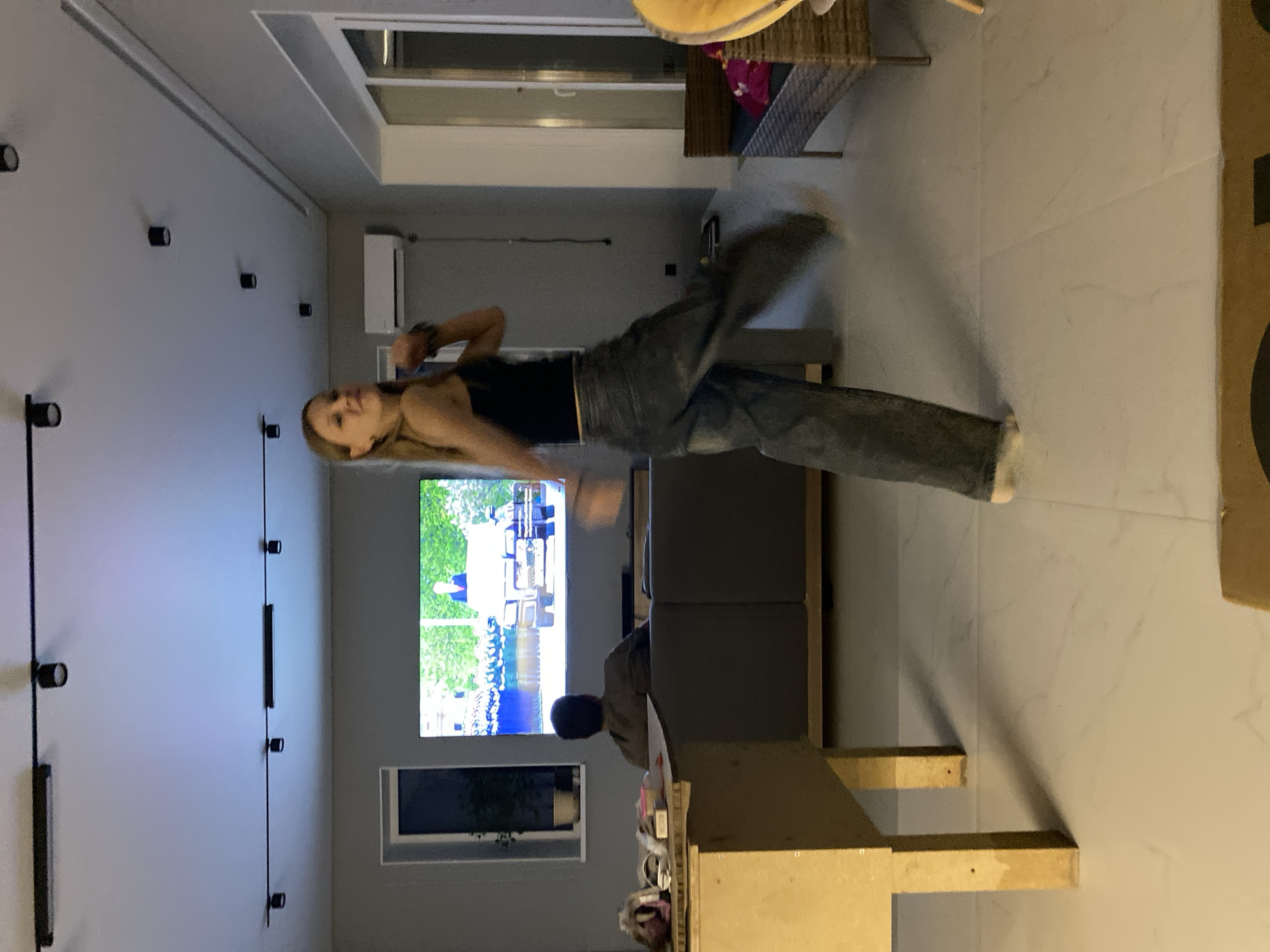 A woman with long blonde hair, wearing a black top and gray jeans, is in mid-air in what appears to be a living room. She is positioned horizontally, with her head towards the left and her feet to the right. A man, likely a child, sits on the edge of a sofa facing the television. The television is displaying a program or video with images of a military parade. The room has white walls with recessed lighting, and the window is at the bottom. The location is unknown; the city of Chaikovsky, Russia is not confirmed by visual evidence.