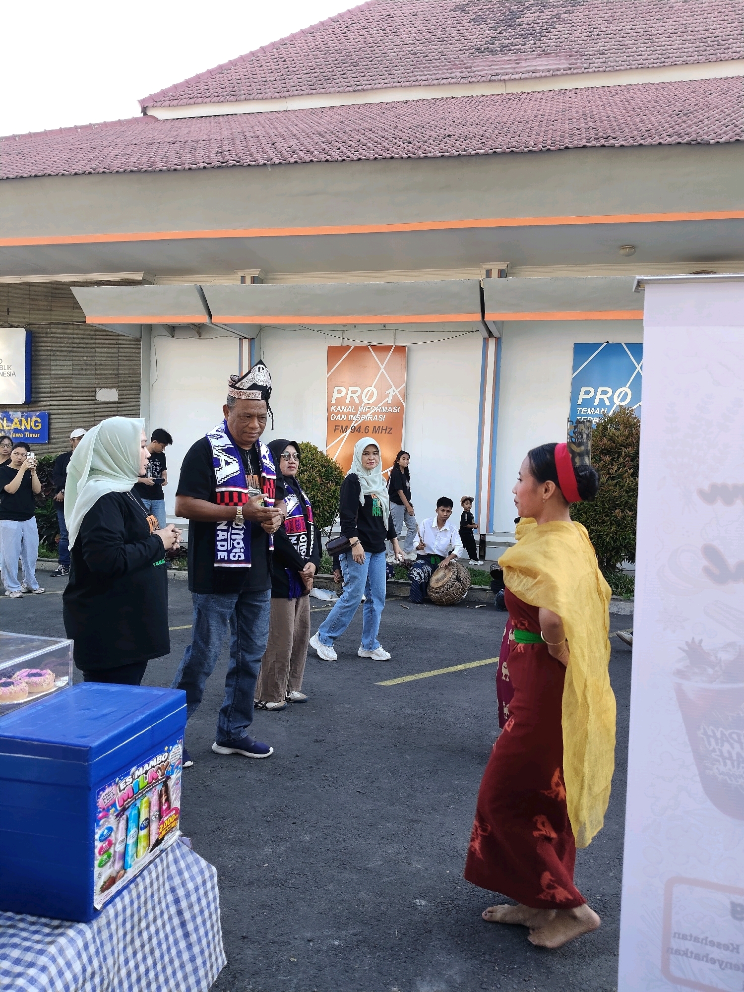 In the city of Malang, Indonesia, an outdoor event is taking place in front of a building with a red tile roof. The ground is asphalt. In the foreground, to the left, is a blue cooler on a table covered with a blue and white checkered tablecloth. The cooler has colorful packaging for "ES MAMBO MILKY." Next to the cooler, in a clear display case, are two pink donuts.
Several people are gathered in the middle ground. A man in a black shirt, blue jeans, and a decorative headdress and sash is speaking to a woman in a black shirt and hijab. Behind them, other individuals are standing and observing. To the right, a young woman in traditional Indonesian attire, including a maroon dress with intricate patterns and a yellow sash, stands barefoot facing away from the viewer. She has a red headband in her hair, which is styled in a bun. Behind her, another young woman in similar traditional dress is partially visible. Further in the background, some people are seated, and one person is playing a drum.
The building behind the participants has a gray facade with orange trim. Several large banners with the "PRO" logo and text are displayed on the building's exterior. One banner is blue and white, while another is orange and white. The event appears to be an outdoor cultural gathering or market.