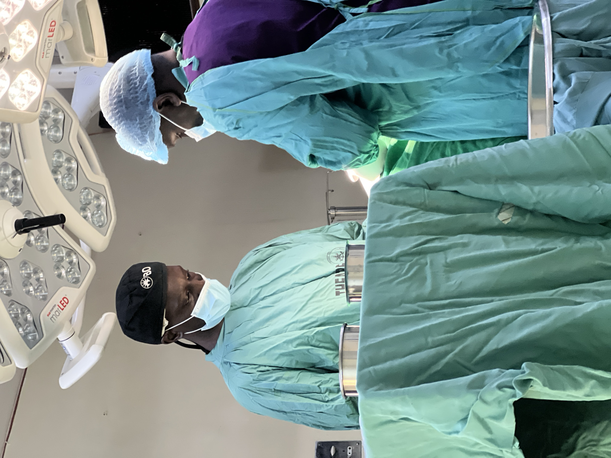 Two medical professionals in surgical attire stand in what appears to be an operating room. They are wearing surgical caps, masks, and gowns in green and blue hues. One individual is wearing a black cap with the letters "BO" on it. The setting includes surgical lights and what appears to be a surgical table covered with green drapes. It is not possible to confirm the location in the city of Nairobi, Kenya based on the visual information provided.