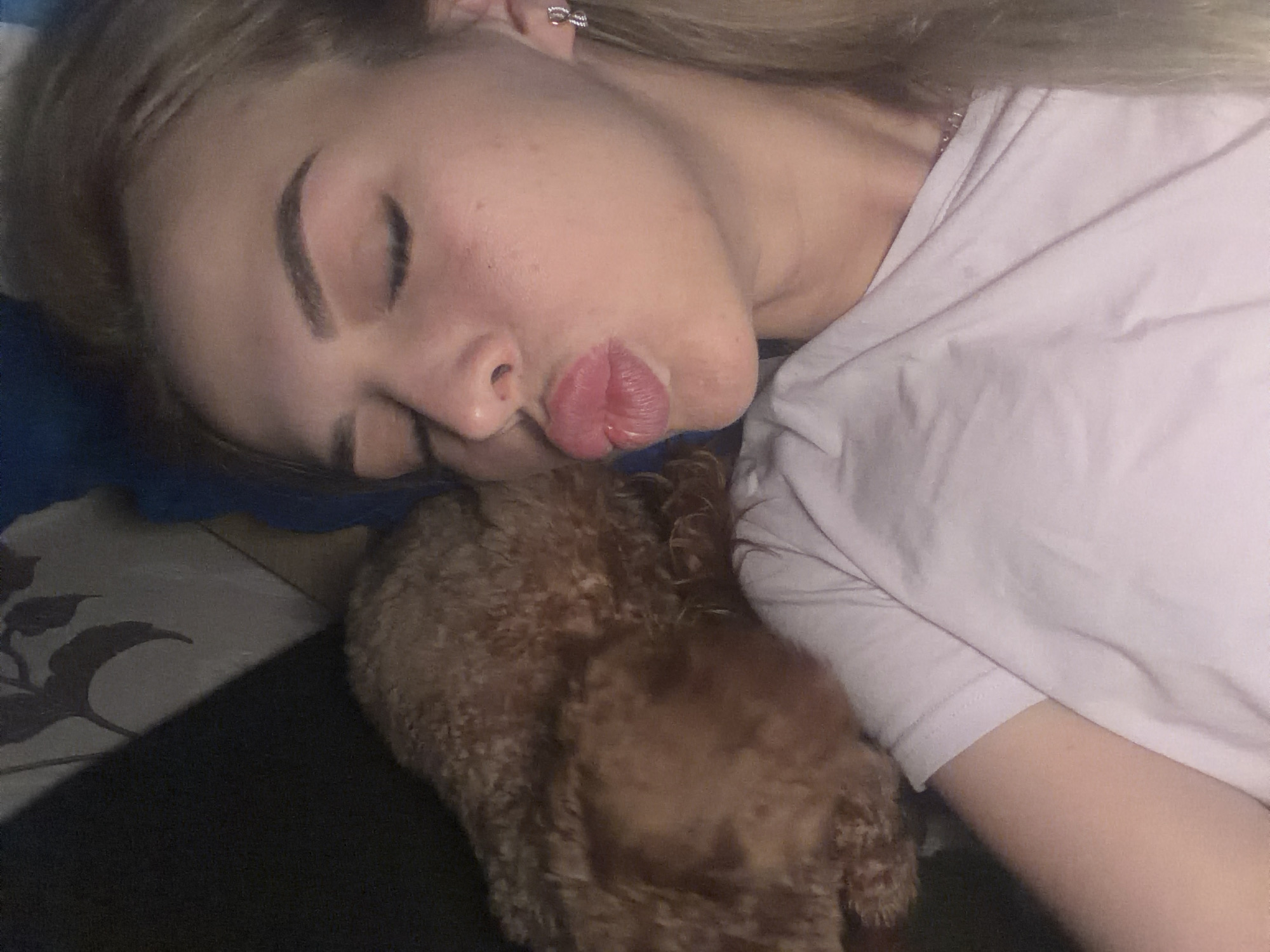 In a dimly lit, indoor setting, a young woman with blonde hair is captured in a close-up selfie. She is wearing a light-colored t-shirt and has her eyes closed, puckering her lips as if for a kiss. A fluffy, light brown dog is nestled beside her, partially obscuring her left shoulder and upper body. The background appears to be a bed or sofa with a patterned fabric visible behind the woman's head, featuring a dark, abstract floral design. The overall ambiance is intimate and casual, suggesting a moment of affection between the woman and her pet. There is no indication of the specific location in Kyiv, Ukraine.