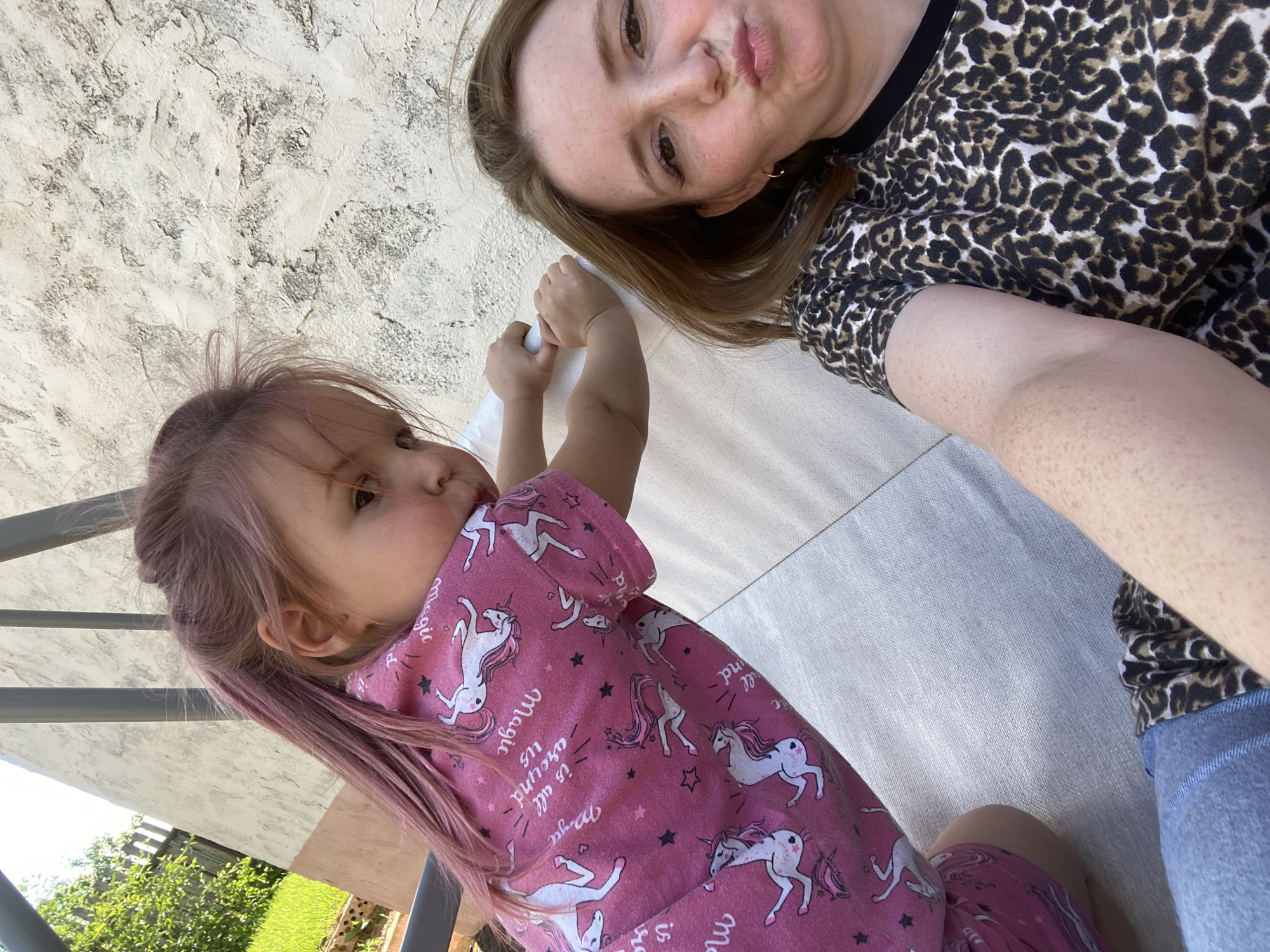In a close-up, selfie-style shot, a woman with auburn hair and freckles pouts at the camera, her head tilted slightly. She is wearing a black and brown leopard print t-shirt. To her left, a young girl with light brown hair and a pink unicorn-print t-shirt leans forward, her gaze directed away from the camera. Her hands are resting on a light-colored cushioned surface. The background shows a textured, light-colored wall and some greenery. The scene appears to be outdoors, likely in a backyard or patio area. No specific location in Chaikovsky, Russia, can be identified from the image.