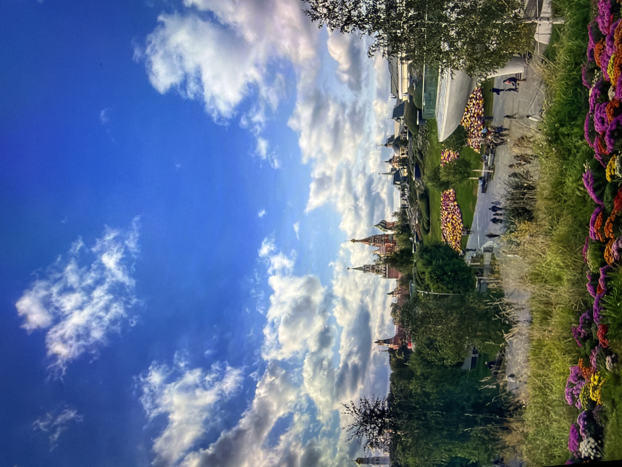 The image depicts an outdoor scene with a prominent view of the Kotelniki Kremlin in the background, characterized by its red brick walls and distinctive onion-domed cathedrals. In the foreground, a lush landscape features a variety of trees, tall grasses, and a vibrant display of colorful flowers, predominantly in shades of purple, pink, yellow, and orange. A paved walkway winds through the scene, where several individuals are walking and sitting on benches. A modern, curved white structure, possibly a building or part of a park installation, is visible on the right side of the image. The sky above is a clear blue, punctuated by fluffy white clouds.