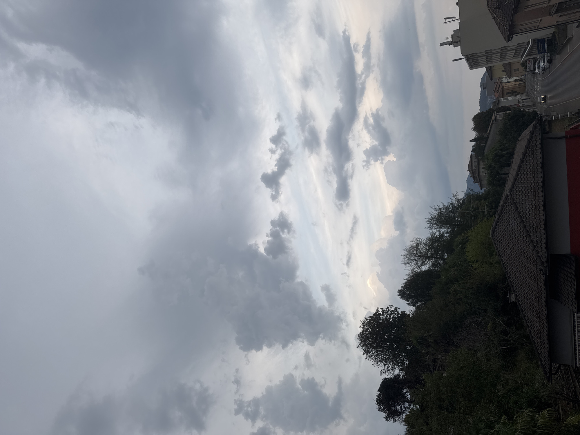The image depicts an overcast sky with dramatic cloud formations over a hillside dotted with trees and buildings. In the foreground, the roof of a building with dark tiles is visible. Beyond this, a densely wooded hillside rises, with glimpses of structures within the foliage. Further in the distance, a street winds through a town, lined with buildings of varying architectural styles, including a modern multi-story structure with antennas on its roof. A few cars are present on the street, with one car's headlights illuminated. The overall impression is of a somewhat moody or pre-storm atmosphere in an urban or semi-urban setting.