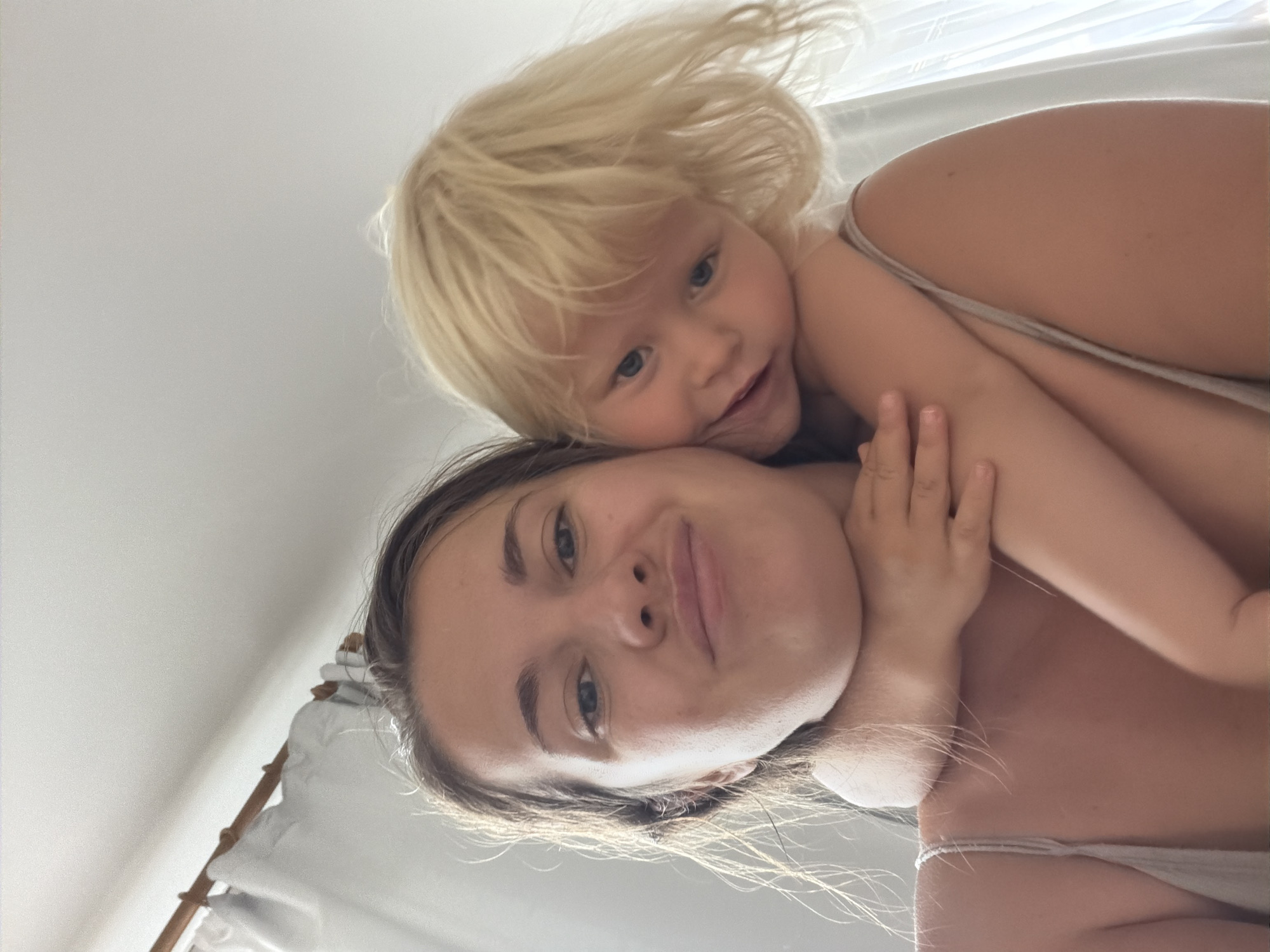 This media file is a close-up, low-angle selfie of a woman and a young child. The woman is in the foreground, looking up at the camera with a slight smile. Her dark hair is visible, framing her face. The child, who appears to be a toddler with blonde hair, is behind and above the woman, hugging her. The child is also looking towards the camera with a happy expression. Both subjects are wearing light-colored tank tops or similar casual clothing.
The scene appears to be indoors, with a bright, diffused light source coming from above and to the right, suggesting daytime. The background is a plain, light-colored wall or ceiling, with some soft fabric visible on the left, possibly curtains or bedding, and what looks like a wooden structure with some white fabric draped over it, which could be part of a crib or a tent.
The overall mood is affectionate and intimate, capturing a tender moment between a mother and child. There is no visible text in the image. The lighting suggests a bright day, possibly morning or afternoon. Given the location context of Sochi, Russia, the setting might be a home or a hotel room.