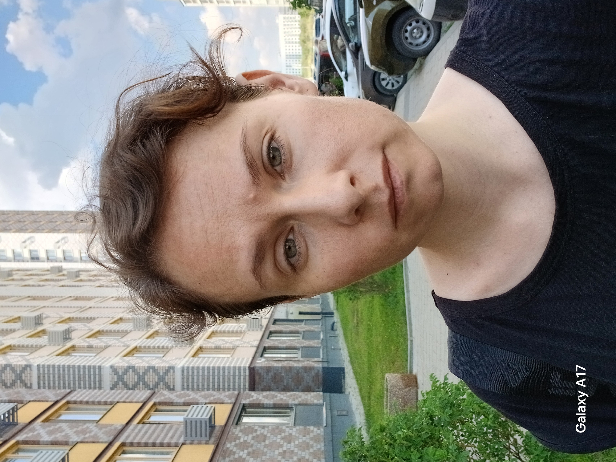 The image shows a woman with short, light brown hair and green eyes. She is wearing a black t-shirt. The background shows a building with a patterned brick facade and yellow accents. There is also greenery and parked cars visible. The camera used to take the image is a "Galaxy A17". Based on the architecture and the location of the image, it is likely taken in Yekaterinburg, Russia.