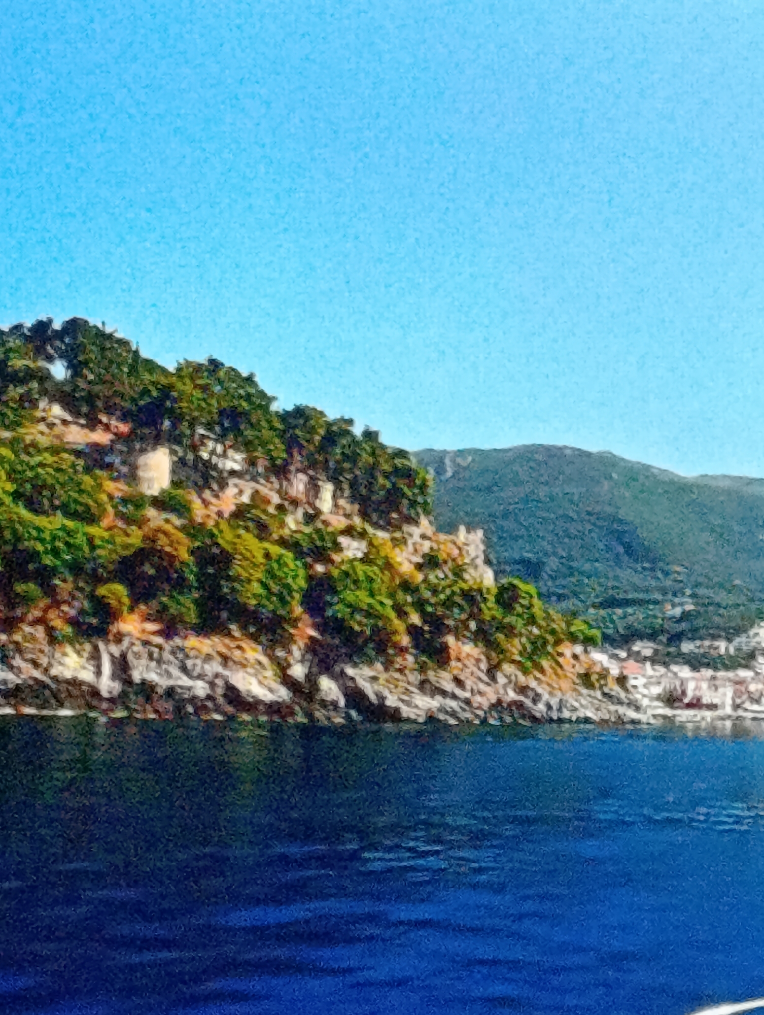 The image shows a coastal scene with a rocky coastline covered in vegetation, possibly trees and shrubs. Above the foliage, there's a structure resembling a castle or fortress. In the background, a hill or mountain is visible. To the right of the coastline, there's a town with buildings clustered near the shoreline. The foreground is dominated by the dark blue water of the sea. The sky is clear and blue. The location is likely a coastal area, possibly somewhere like the Amalfi Coast.