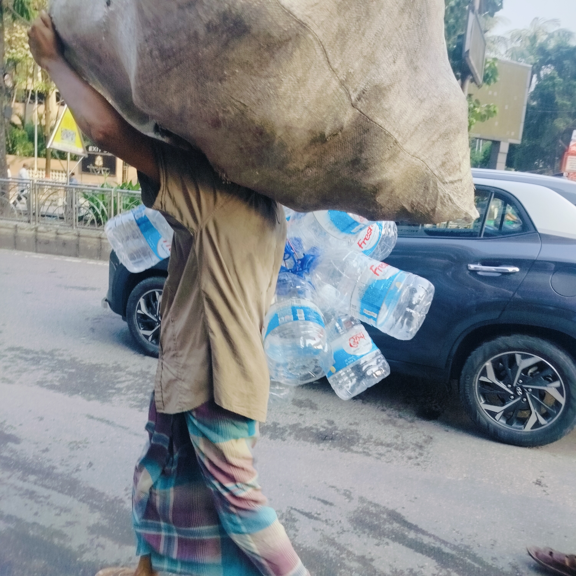 A man is walking on a paved road, carrying a large sack over his head and a bunch of water bottles in his right hand. He is wearing a beige shirt and a colorful, patterned lungi (a traditional garment worn around the waist). The water bottles are labeled "Fresh."
A dark-colored car is parked on the side of the road. The background is blurred, but there are hints of buildings and street signs. The location is likely Dhaka, Bangladesh, based on the common practice of carrying heavy loads in this manner and the presence of lungi.