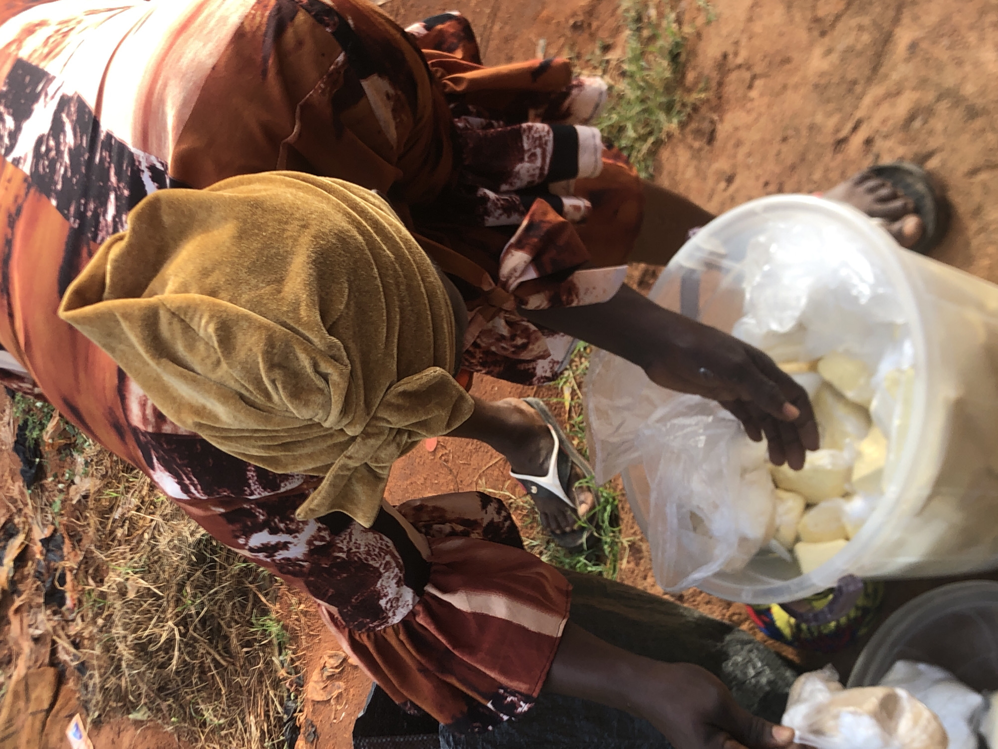 A person wearing a brown and white patterned dress, a brown headwrap, and black and white sandals is outdoors on reddish-brown soil mixed with dry grass, possibly in Ukwu-Abwa, Nigeria. The individual is reaching into a large, translucent plastic container filled with off-white, irregularly shaped solids, possibly shea butter. A smaller, partly filled translucent plastic container sits nearby. The person's skin tone is dark. The individual is in the process of taking some of the solids out of the plastic container to put into the smaller one.