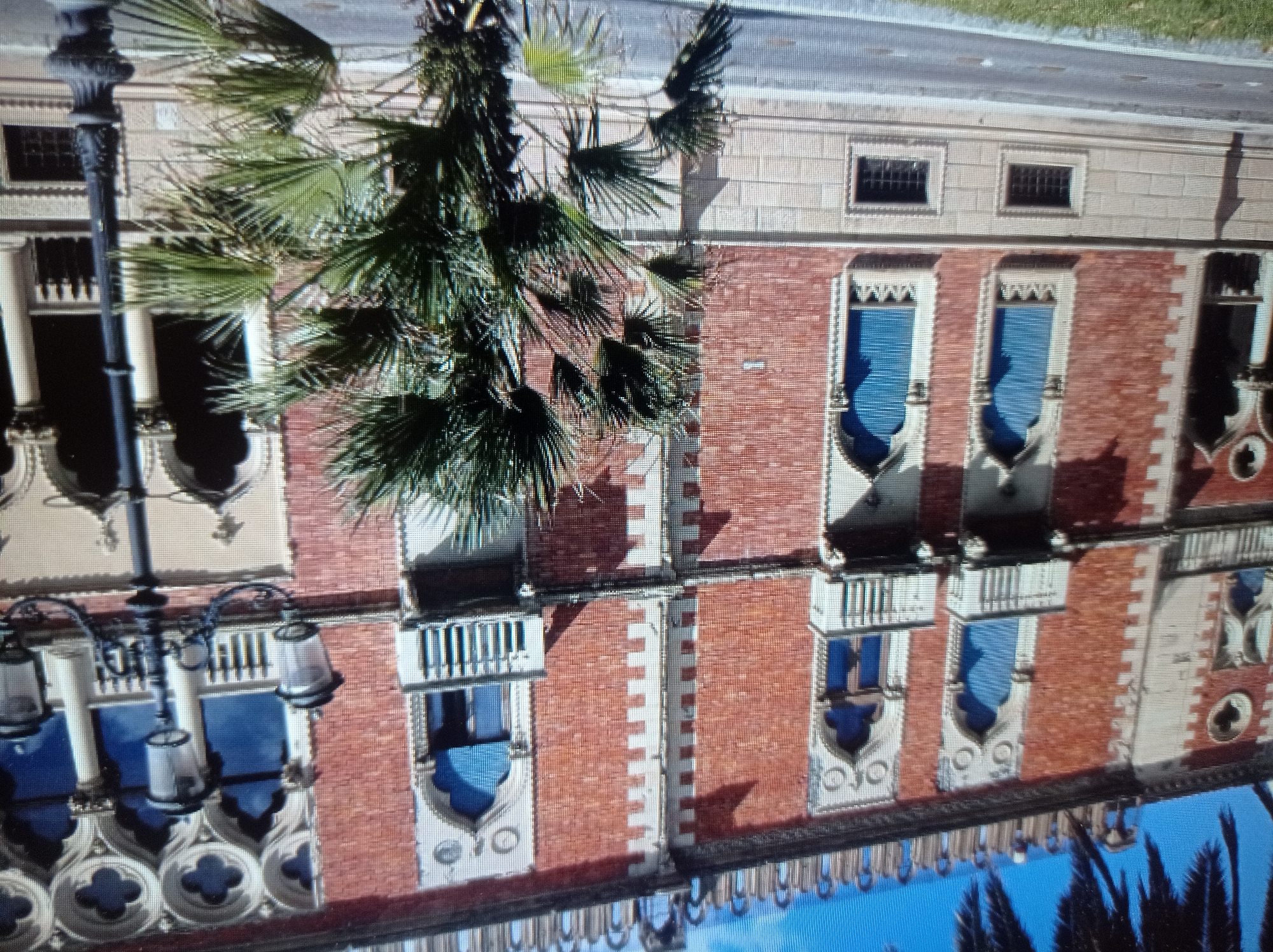 This image depicts a red brick building with ornate white architectural details. The building has arched windows with blue shutters and balconies with decorative railings. A lamppost with multiple lights stands in front of the building. Palm trees are visible in the foreground, partially obscuring the lower part of the building. The scene appears to be a street view in a city. The location in the city of Mukachevo, Ukraine is not identifiable from the visual information presented in the image.