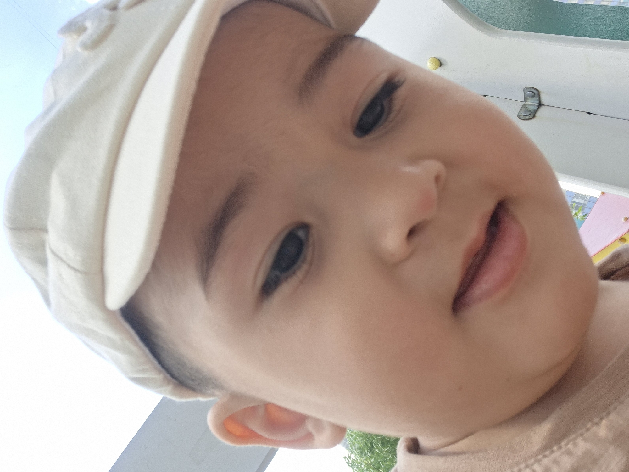 A close-up, low-angle portrait features a young child, possibly of East Asian descent, wearing a light-colored fabric cap and a light brown t-shirt. The child's face is prominent, with dark eyes looking slightly downwards and a partially open mouth revealing the tip of the tongue. Short, dark hair is visible beneath the cap, particularly above the left ear. The upper background shows a bright, light blue or overcast sky. To the left, a light-colored architectural element and green foliage are partially visible. The right and lower-right background features a white structure, indicative of playground equipment, with details including a yellow spherical cap or bolt, a metallic clasp or hinge, and a teal-colored panel beyond. The scene is located in the city of Unknown, Russia.