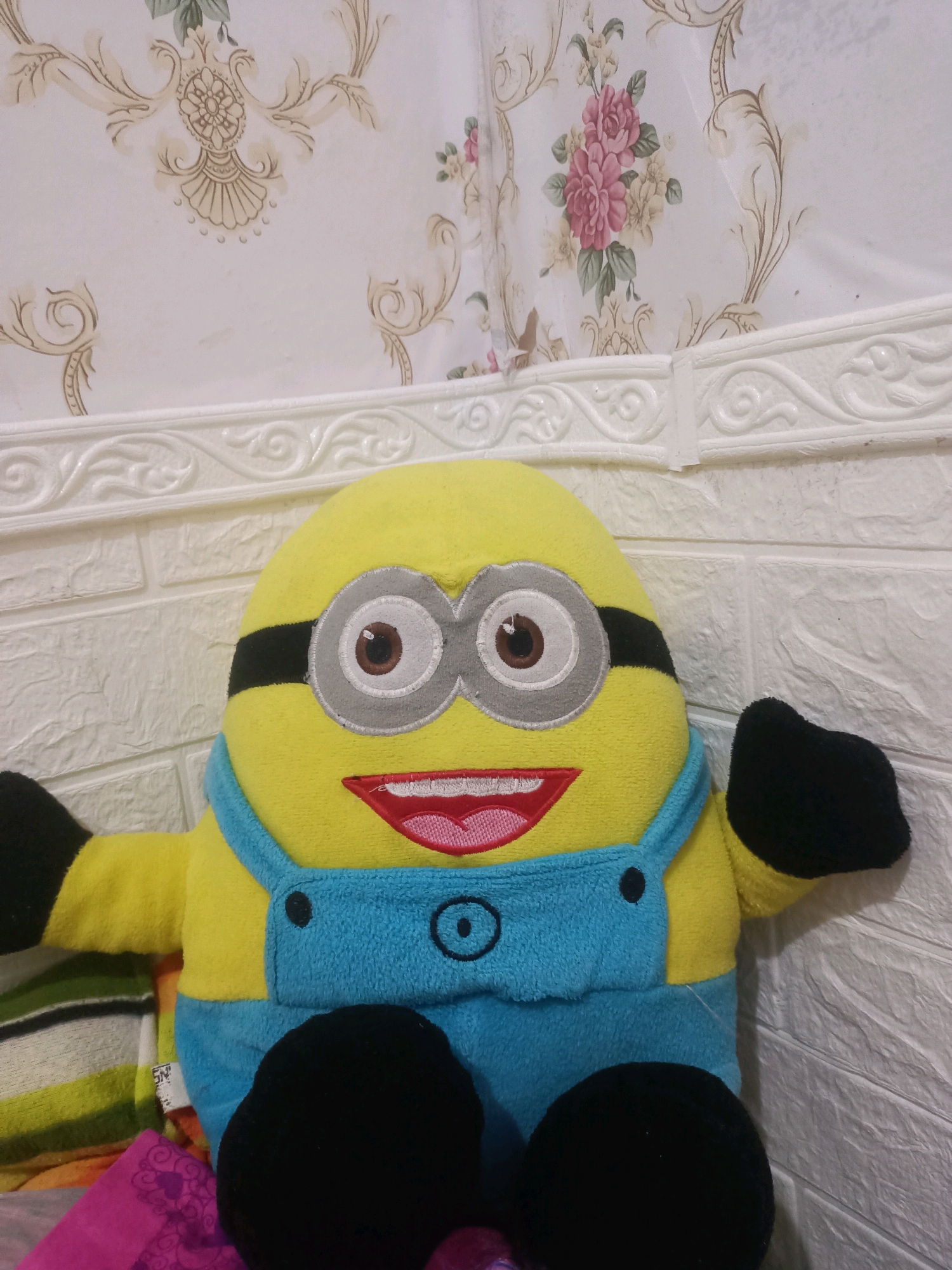 A plush toy, resembling a Minion character from the movie "Despicable Me," is the central focus of the image. The toy is predominantly yellow with blue overalls and black goggles. It is positioned in the foreground, with its arms outstretched and legs visible. Behind the toy, a white brick-patterned wall serves as the background, and above it, a patterned wallpaper with floral designs is displayed. The lighting appears to be artificial, casting soft shadows. The overall scene suggests an indoor setting, likely a room in Wadeng, Indonesia.