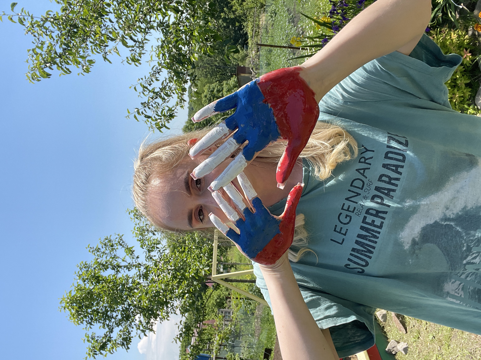 In the city of Yasnoe, Russia, a young woman with blonde hair stands in a garden on a sunny day. She is wearing a teal t-shirt with the text "LEGENDARY SUMMER PARADISE RELAX & SURF" printed on it. Her hands are painted with the colors of the Russian flag: red on the palm and lower fingers, blue in the middle, and white on the fingertips. She is holding her hands up in front of her face, partially obscuring it, as she looks at the camera. The background shows lush green trees, a fence, and a clear blue sky, suggesting an outdoor recreational area.