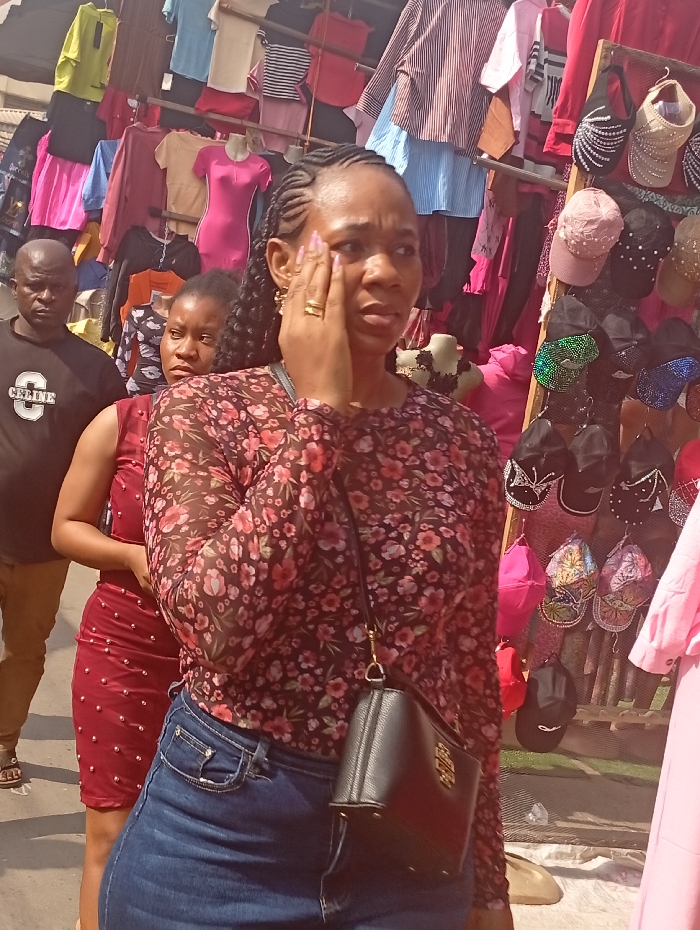 The scene depicts a busy outdoor market environment in Fegge, Nigeria. In the foreground, an adult female of African descent is centrally positioned, looking slightly to her left with her right hand touching the right side of her face. She has braided hair, wears a dark long-sleeved blouse with a red and pink floral pattern, blue denim jeans, and a black textured cross-body bag with a gold-colored accent. A gold-colored ring is visible on her right ring finger.
Behind her, to the left, another adult female of African descent is partially visible, wearing a maroon-colored dress adorned with metallic studs, looking towards the left. Further behind her, to the far left, an adult male of African descent, bald, is partially visible, wearing a dark t-shirt with a white "CELINE" logo and light-colored trousers, also looking left.
The background is filled with various clothing items displayed for sale. Multiple brightly colored garments, including pink dresses, striped shirts, and various tops, hang on racks and mannequins. To the right, a vertical display stand holds an assortment of caps and visors, many of which are embellished with rhinestones or sequins in colors such as pink, black, and iridescent green. The lighting is bright, indicating daylight.