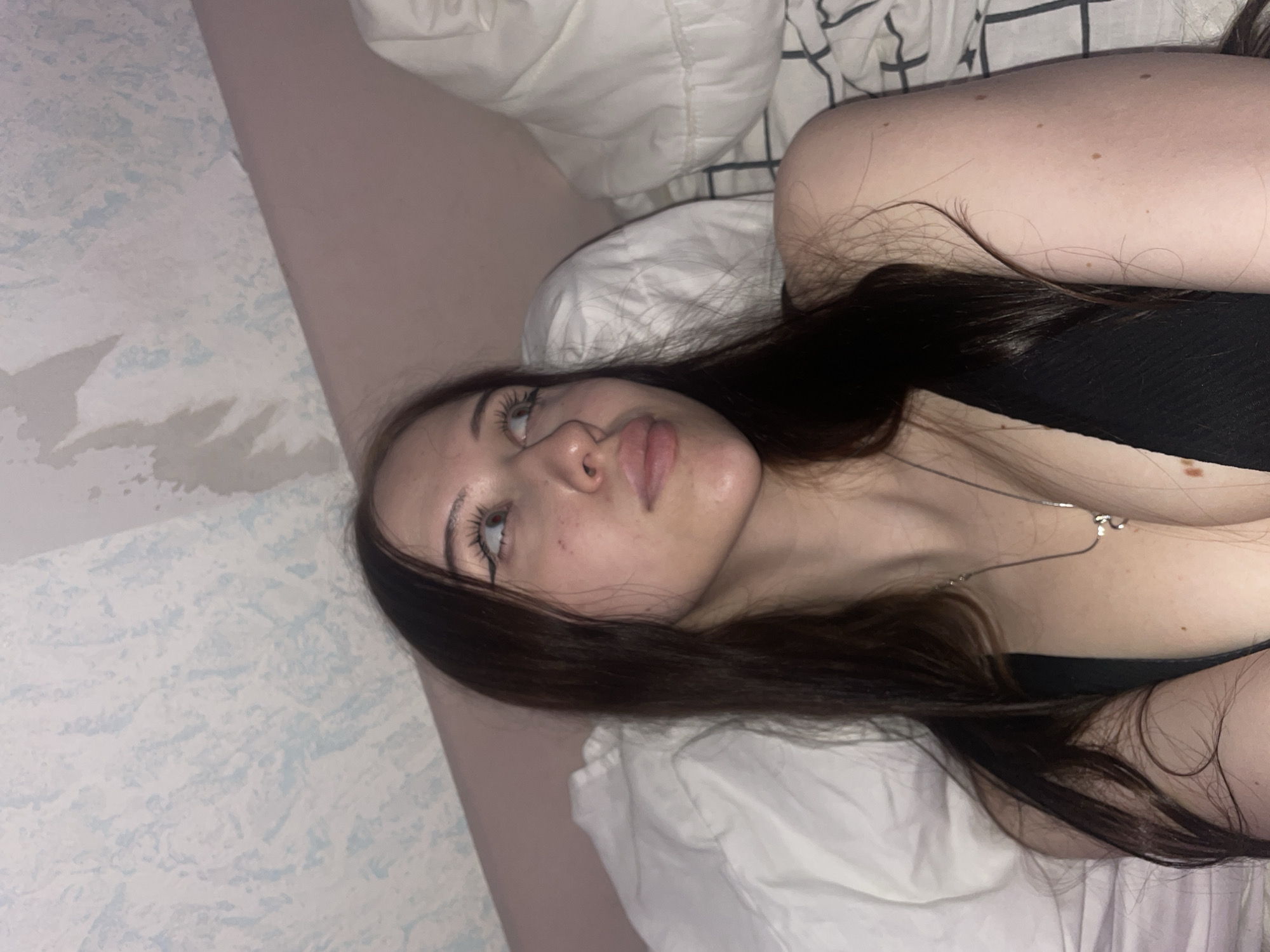 In a close-up, indoor shot taken in Votkinsk, Russia, a young woman with long, dark hair lies on a bed. She is wearing a black top and a silver necklace. Her gaze is directed upwards and to the right, with her head tilted slightly. The background consists of pillows and a wall with a textured, light-colored wallpaper. The lighting is dim, suggesting an indoor environment, possibly a bedroom.