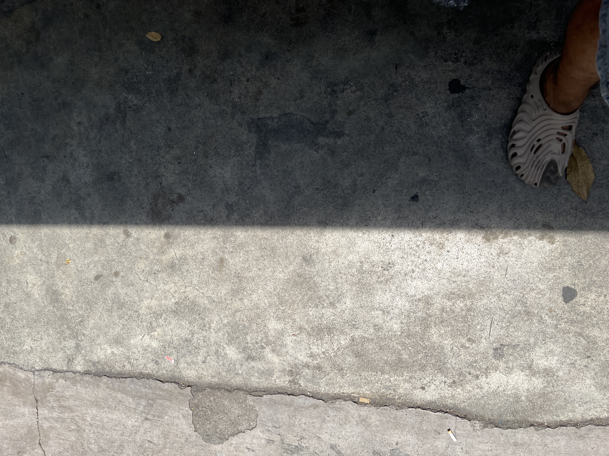 The image depicts a section of a concrete sidewalk in Nha Trang Ward, Vietnam. The sidewalk is bisected by a sharp shadow, casting half of it into darkness. On the sunlit side, the concrete is cracked and worn, with a single cigarette butt visible near the edge. A small piece of red paper is also present. The shaded side of the sidewalk is darker and appears smoother, with a single dry leaf lying on it. In the lower right corner, a person's foot wearing a light-colored, avant-garde sandal is visible, stepping near another dry leaf. The scene suggests an urban outdoor environment, possibly a street or pathway.