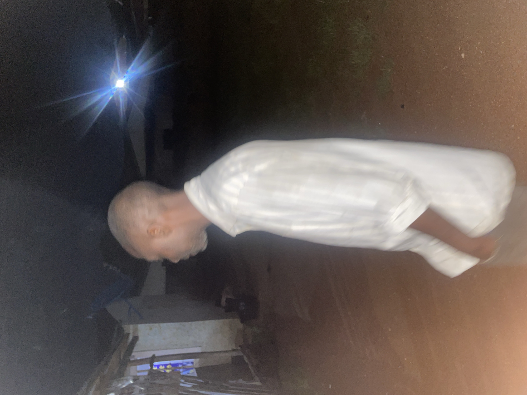 The image depicts a dimly lit outdoor scene, likely at night, with a man in the foreground. The man is wearing a light-colored, possibly white, striped shirt and appears to be walking away from the camera, facing left. His skin tone is dark, and he has short, graying hair and a beard. His expression is not visible.
Behind the man, the environment is dark and indistinct. To the left, there appears to be a structure with a roof, possibly a building or shelter, with some light emanating from within. The ground is a reddish-brown, suggesting it might be dirt or unpaved earth. In the background, a bright light source with prominent starburst effects illuminates the sky, suggesting a powerful, distant light, possibly a streetlamp or a floodlight. Some vegetation can be seen on the right side of the image.
While the image itself does not provide explicit clues to the precise location within Jos, Nigeria, the general characteristics of the environment—the unpaved ground, the simple structures, and the outdoor setting—are consistent with many residential or semi-urban areas in the region. The presence of a bright light source in the distance might indicate an area with some infrastructure, even if rudimentary.