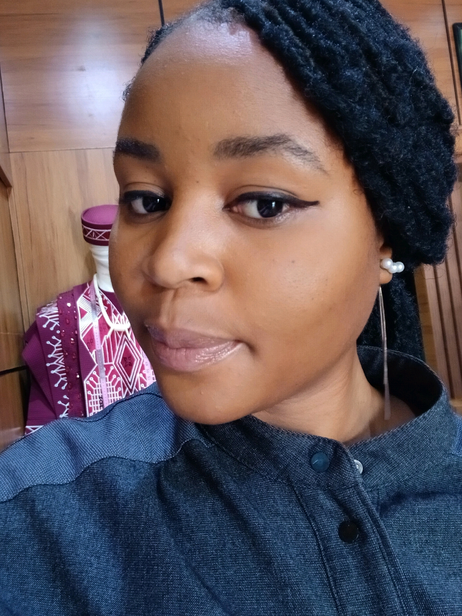 A close-up, head-and-shoulders selfie shows a young Black woman with dark skin and black braided hair. She is looking directly at the camera. She is wearing a dark denim shirt with a collared neckline and visible buttons. She has on makeup, including eyeliner and lip gloss. A long, silver earring with three small pearl-like beads is visible on her right ear. In the background, to the left of the woman, is a mannequin dressed in a purple and pink patterned garment with a matching hat. The background appears to be an indoor setting with wooden paneling on the wall.