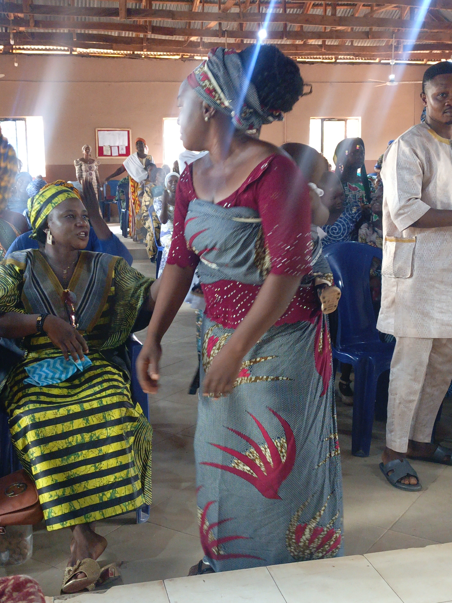 The image captures a scene in Akwanga, Nigeria. A group of people are gathered indoors. A woman in a colorful striped dress and headwrap is seated, holding a light blue cloth. Another woman, wearing a patterned gray wrap dress with red floral details, has a baby secured on her back. A man in a light-colored outfit stands nearby. Additional people are seated or standing in the background, amidst blue plastic chairs. The environment appears to be a communal space with a wooden roof structure.