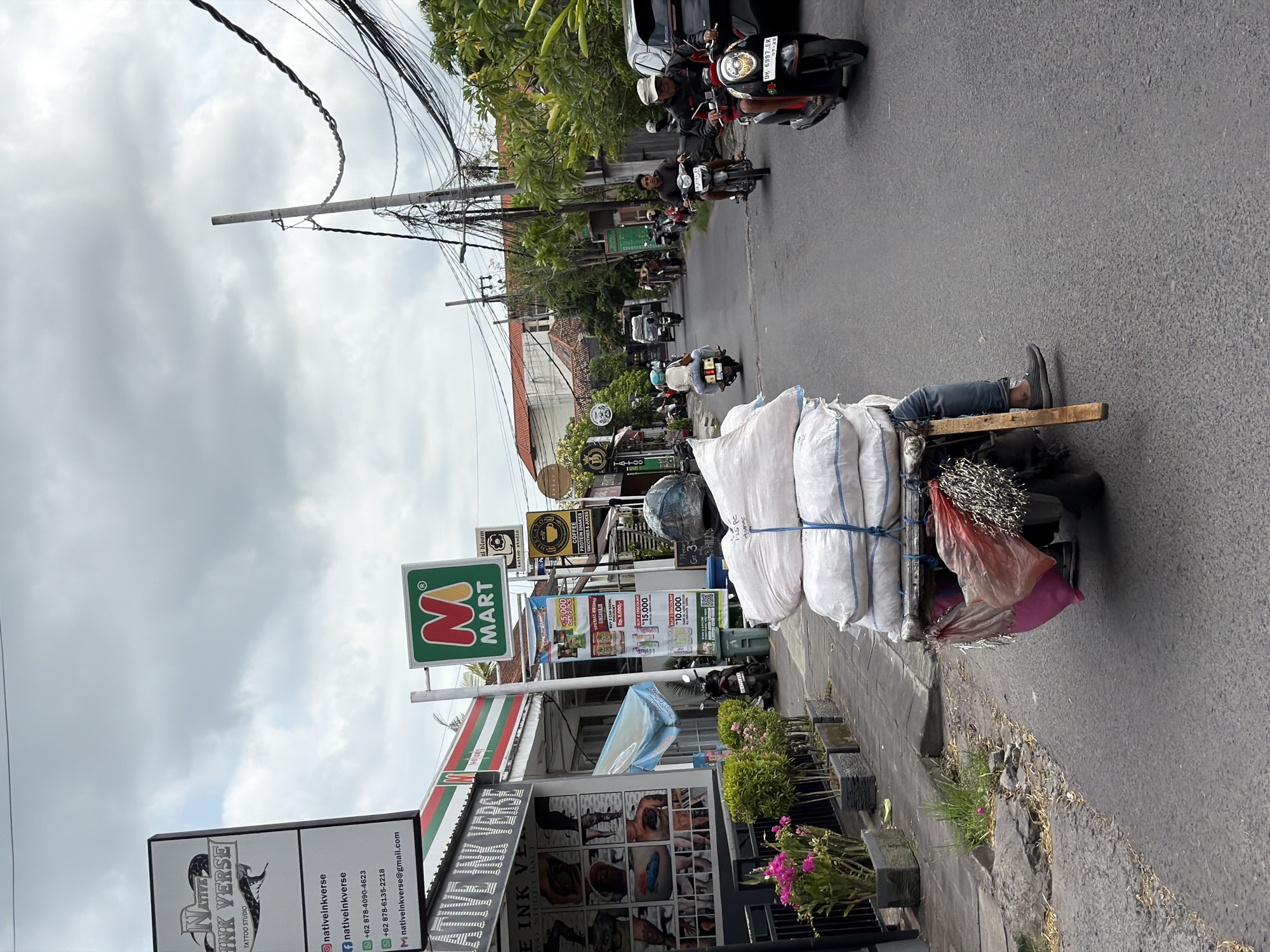 The image depicts a street scene in an urban environment, possibly in Southeast Asia, under an overcast sky. In the foreground, a motorcycle is heavily laden with several large, white sacks tied with blue rope. A person wearing jeans and a helmet is seated on the motorcycle, operating it. The motorcycle's rear is adorned with pink and red plastic bags, and metallic fringe.
To the left of the road, a sidewalk runs alongside shops. One shop prominently displays a sign for "NATIVE INK VERSE TATTOO STUDIO" with images of tattoos on its facade. Adjacent to it is a "M MART" convenience store with its logo visible. Further down the street, more shops and buildings are visible, along with dense foliage and trees.
Several other motorcycles are present on the road. One motorcycle to the right carries a rider wearing a white helmet and dark jacket. Another motorcycle ahead of it has a rider in a dark shirt. In the distance, more vehicles and people can be seen, suggesting moderate traffic. Utility poles with numerous tangled wires line the side of the street. The road surface is asphalt, with some cracks and patches visible. The overall impression is a bustling commercial street with significant motorcycle activity and pedestrian access to shops.