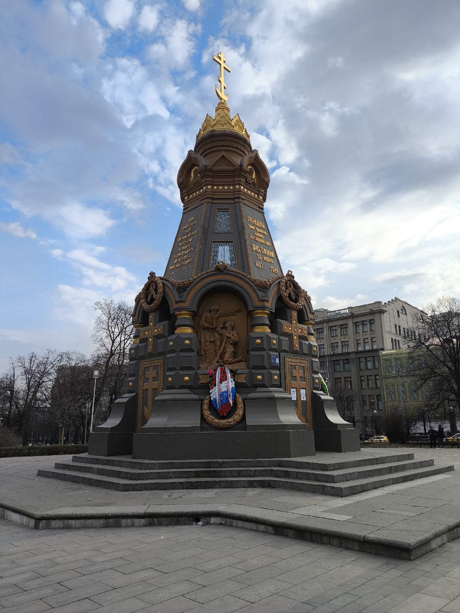 The image shows the Monument to the Heroes of Pleven in Moscow, Russia. The monument is a tall, dark-colored structure with golden accents, topped with a golden cross. It has a bas-relief depicting soldiers, and a wreath with the colors of the Russian flag is placed at its base. The monument is set on a multi-tiered stone platform, surrounded by a gray-paved plaza. In the background, there are bare trees and buildings, with a cloudy sky above. Some people and yellow taxis are visible in the distance.