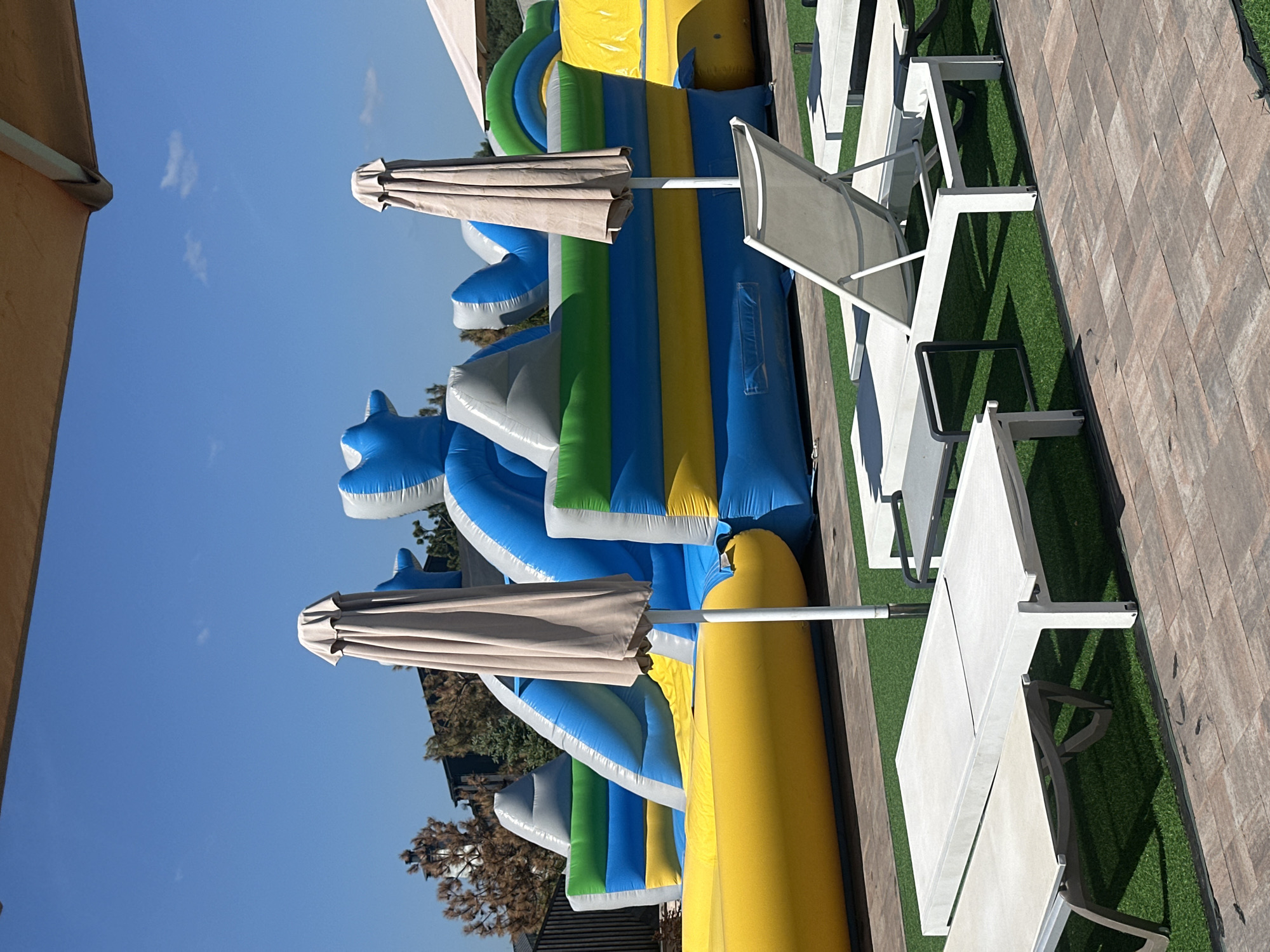 The image shows a sun loungers set up on a green lawn, next to a large inflatable water slide. The water slide is blue, green, and yellow, with a large yellow tube at the bottom. There are two umbrellas open, providing shade for the loungers. The scene is outdoors, with a clear blue sky and some trees visible in the background. The lighting suggests it is daytime, likely mid-morning or afternoon given the shadows. The overall atmosphere appears to be relaxed and recreational, suggesting a resort or a park. There is no visible text in the image.