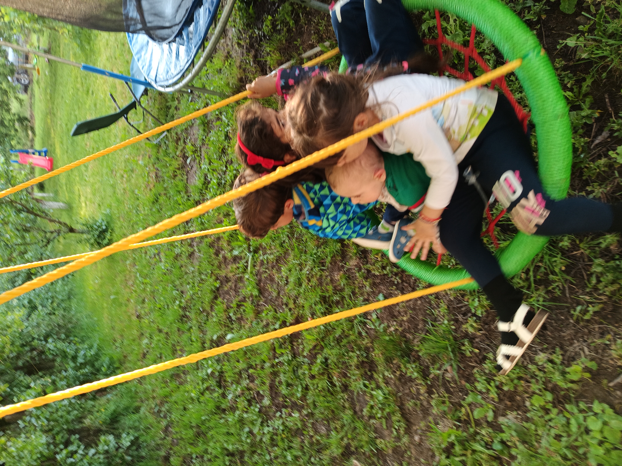 The image depicts an outdoor recreational scene in Kovachevtsi, Bulgaria. Four children are seated within a bright green, circular "nest" or "saucer" swing, which is suspended by multiple yellow ropes visible in the foreground and midground. From left to right within the swing: a child with brown hair wears a blue and green patterned top; behind them, a child with brown hair and a red headband wears a dark top with light patterns; a younger child or infant with lighter hair wears a green top and is nestled close to a fourth child. This fourth child, with brown hair, wears a white long-sleeved shirt, dark blue leggings, and white and brown sandals on the visible foot, appearing to support the younger child. Motion blur is evident, indicating the swing is in motion. The ground beneath the swing consists of grass with patches of exposed soil. In the background, to the right, a blue and black trampoline and a dark green folding chair are visible. Further back, a red and blue children's slide is present. The entire scene is surrounded by green trees and dense foliage under natural daylight.