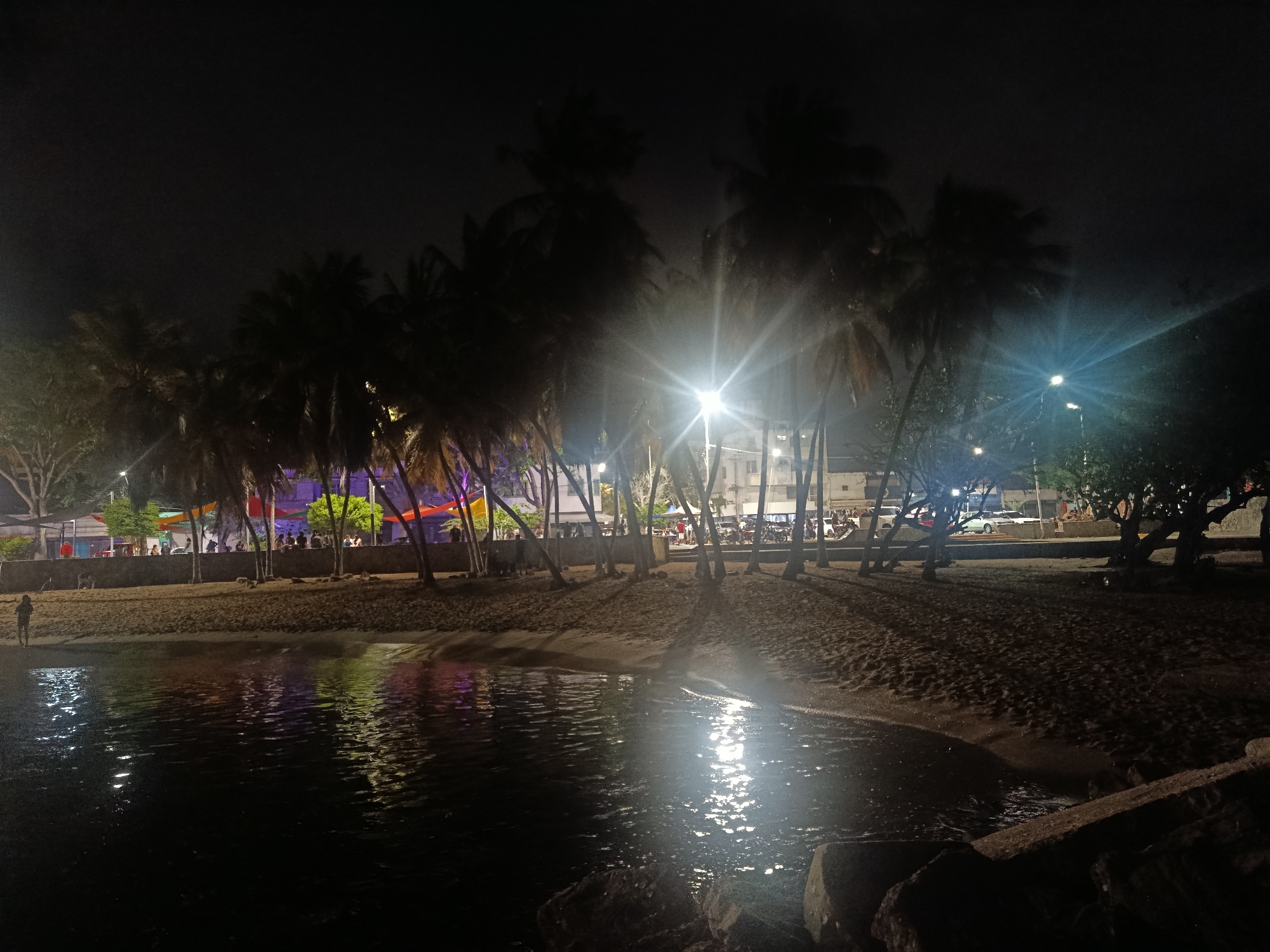 The image depicts a nighttime scene at a beach or waterfront area, likely in Juan Griego, Venezuela. The foreground is dominated by dark water reflecting the lights from the shore, with large rocks visible in the lower right corner. The midground features a sandy beach with palm trees casting shadows. Along the beach, there is a wall or embankment, behind which a gathering of people can be observed. Colorful banners or flags are strung across the area, suggesting a festive or event atmosphere. Buildings and streetlights are visible in the background, indicating the presence of a town or city. Several vehicles are parked along the road bordering the beach. A single person is standing on the sand near the water's edge, facing away from the camera. The overall impression is a lively nocturnal gathering in a coastal urban setting.