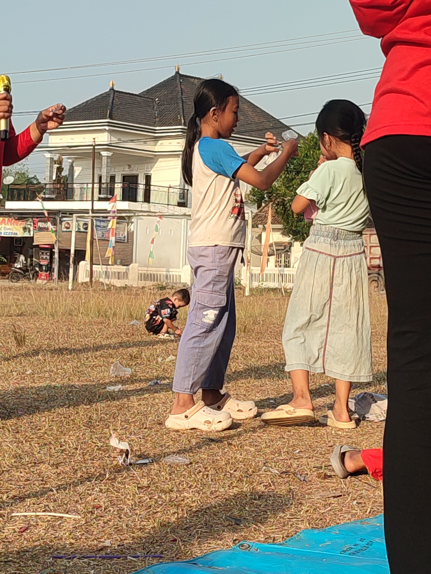 A medium shot captures three children and part of an adult in a sunlit, outdoor setting on the outskirts of Nusa Jaya, Indonesia. The ground is covered in dry grass with scattered debris, suggesting a potentially untidy area.
In the foreground, the lower half of a person in a red shirt and black pants stands on the right, partially obscuring other subjects. To the left, a child wearing a white shirt with blue sleeves and blue pants is holding a small white object, possibly a plastic bottle, and appears to be interacting with another child. This second child, dressed in a light green shirt and a light blue skirt, faces away from the camera, holding a pink object against their chest.
A third child, in the middle ground, is crouched on the grass, facing away from the camera and engaged with something on the ground.
In the background, a multi-story building with a dark, tiled roof and white walls stands prominently. The building has several windows and decorative elements. In front of the building, there are several stalls or small shops, with motorcycles parked nearby, indicating a populated area. Small colorful flags are strung between some of the structures. The sky is clear and light blue, suggesting a sunny day.