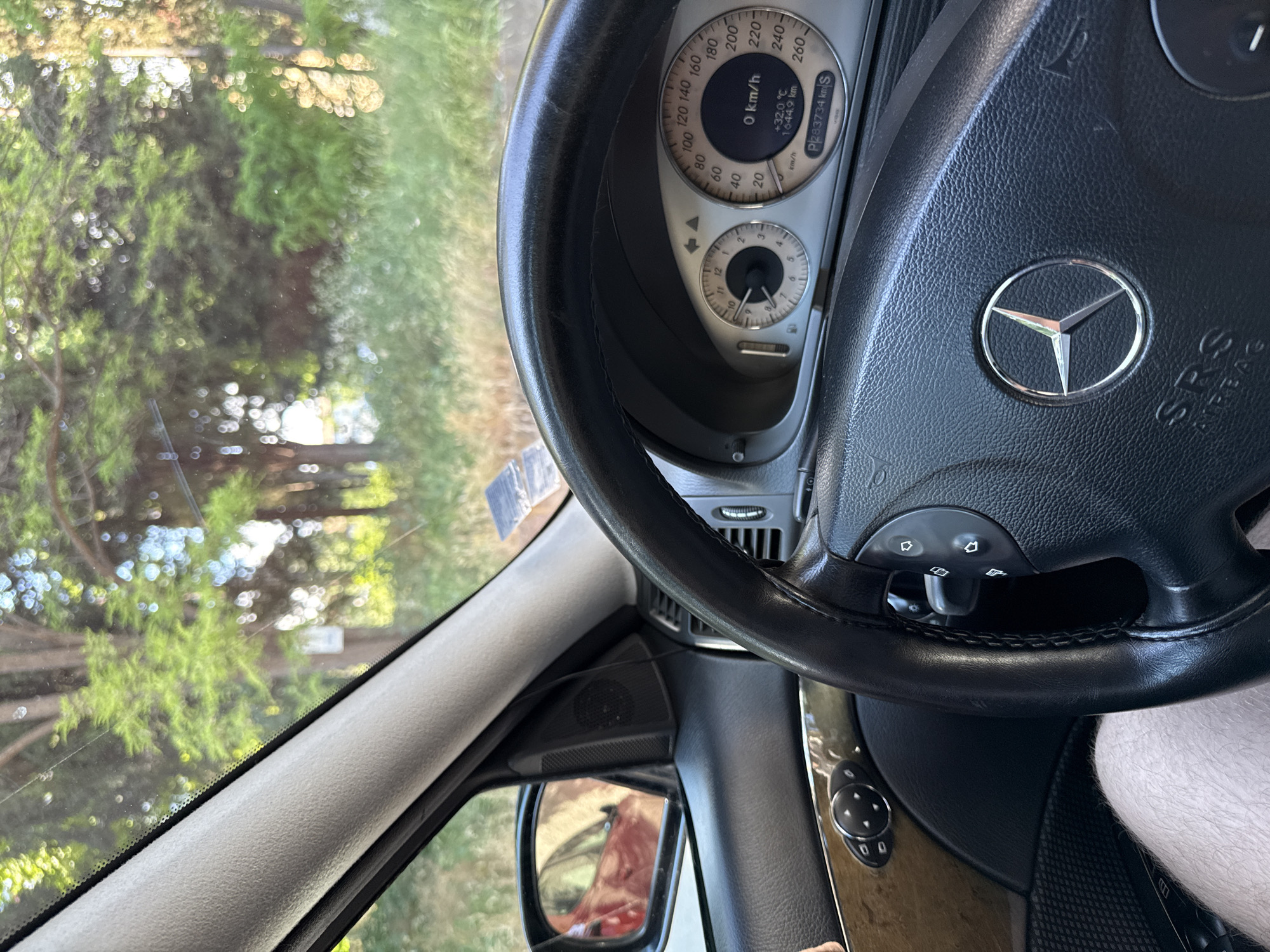 The image shows the interior of a Mercedes-Benz car. The steering wheel with the Mercedes-Benz logo is prominently visible. The dashboard displays a speedometer indicating 0 km/h, an outside temperature of +32.0 °C, and a total mileage of 1644.9 km. The total mileage reading is P283734 km. Through the car window, trees and foliage are visible. The location cannot be determined as Stara Zagora, Bulgaria.