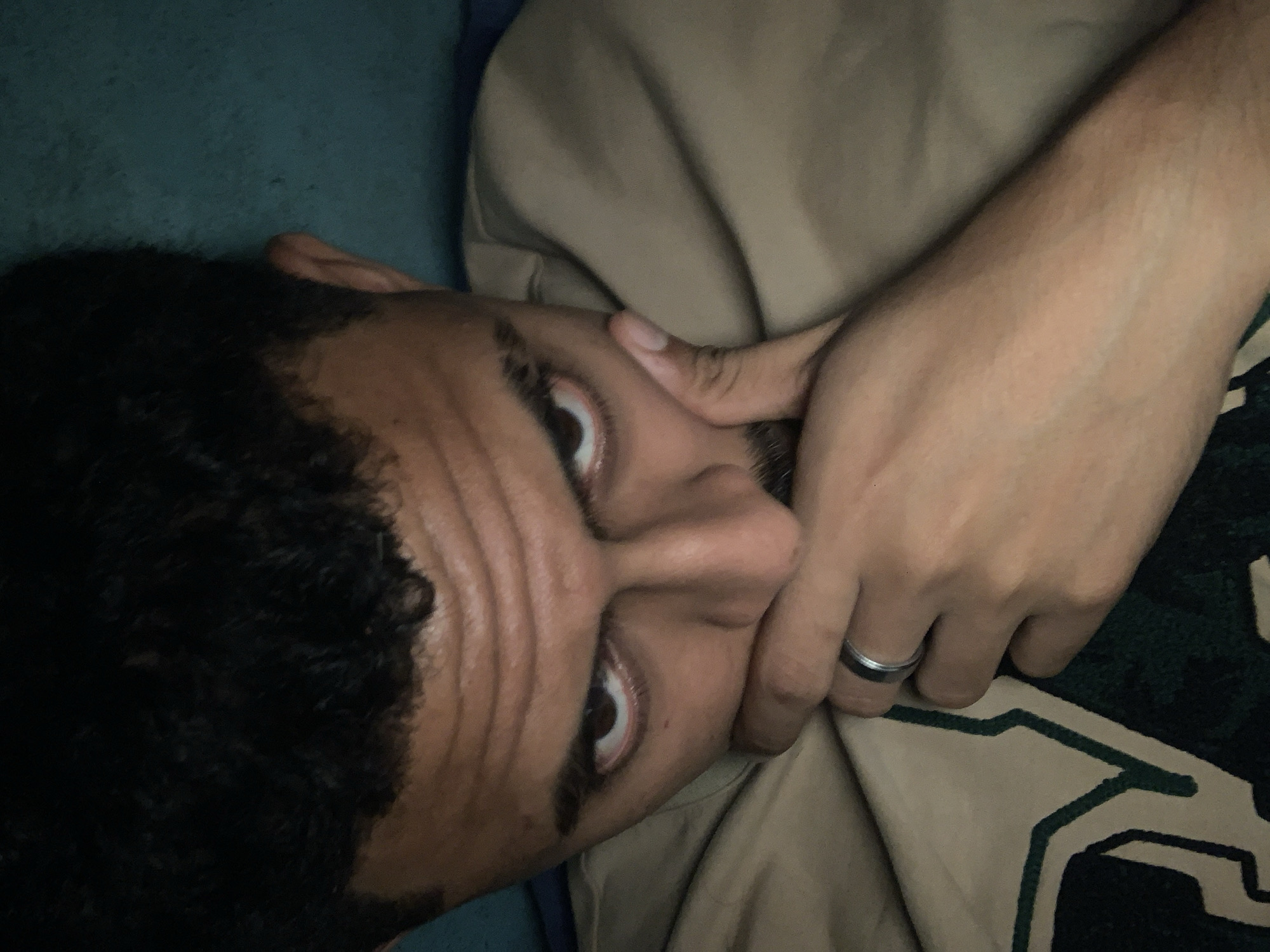 This image captures a close-up, rotated view of a person lying down, with their head positioned towards the left. The subject has brown skin and dark, tightly-curled hair. Their wide-open eyes, looking directly at the viewer, convey an intense, possibly surprised or focused expression, accentuated by several wrinkles across their forehead. One hand, adorned with a silver-toned ring on the ring finger, covers the lower part of their face, from the nose down to the chin.
The person is wearing a light tan or beige garment, which appears soft and draped, likely a shirt or hoodie. Partially visible in the lower right corner is a dark green fabric with a distinctive, lighter geometric pattern, possibly a blanket or part of another piece of clothing. The background behind the subject's head and upper body is a solid, dark teal or blue-green color, suggesting soft furnishings like a pillow or mattress.
The setting is an indoor space, likely a bedroom, given the intimate nature of the shot and the soft fabrics. The lighting is subdued, creating a somewhat dim atmosphere. There is no visible text in the image. The person's gesture of covering their face while maintaining eye contact suggests a moment of introspection, shyness, or a subtle reaction to an unseen event. This scene takes place in Santa Ana, Venezuela.