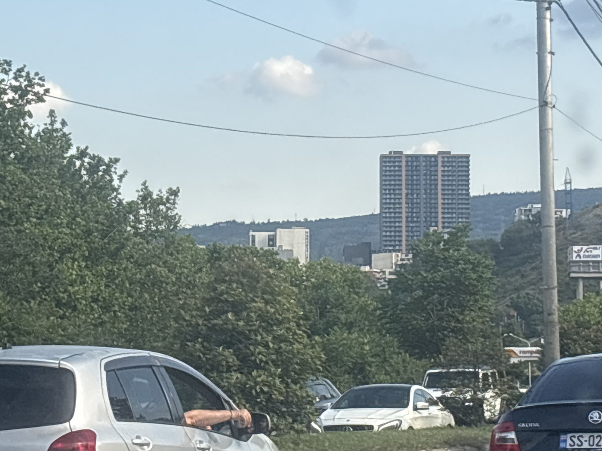The image shows a street scene in Tbilisi, Georgia, likely during the daytime under a clear sky with some scattered clouds. The foreground is dominated by the side view of a silver car, with a person's arm visible resting on the open window. The car is in traffic, as suggested by the presence of other vehicles, including a white Mercedes-Benz in the middle ground.
Behind the cars and a dense line of green trees, a cityscape unfolds. Prominent features include two tall, modern apartment buildings with many windows, suggesting a residential area. Further in the background, a hilly landscape is visible, with more buildings and some communication towers or poles. A large billboard with red and white text, possibly advertising, is also visible on the right side of the image. The overall impression is of a bustling urban environment integrated with nature, characterized by a mix of old and new architecture. The weather appears fair, and the lighting indicates it's daytime, possibly late morning or early afternoon. No specific event is clearly depicted, but the traffic suggests normal daily activity.