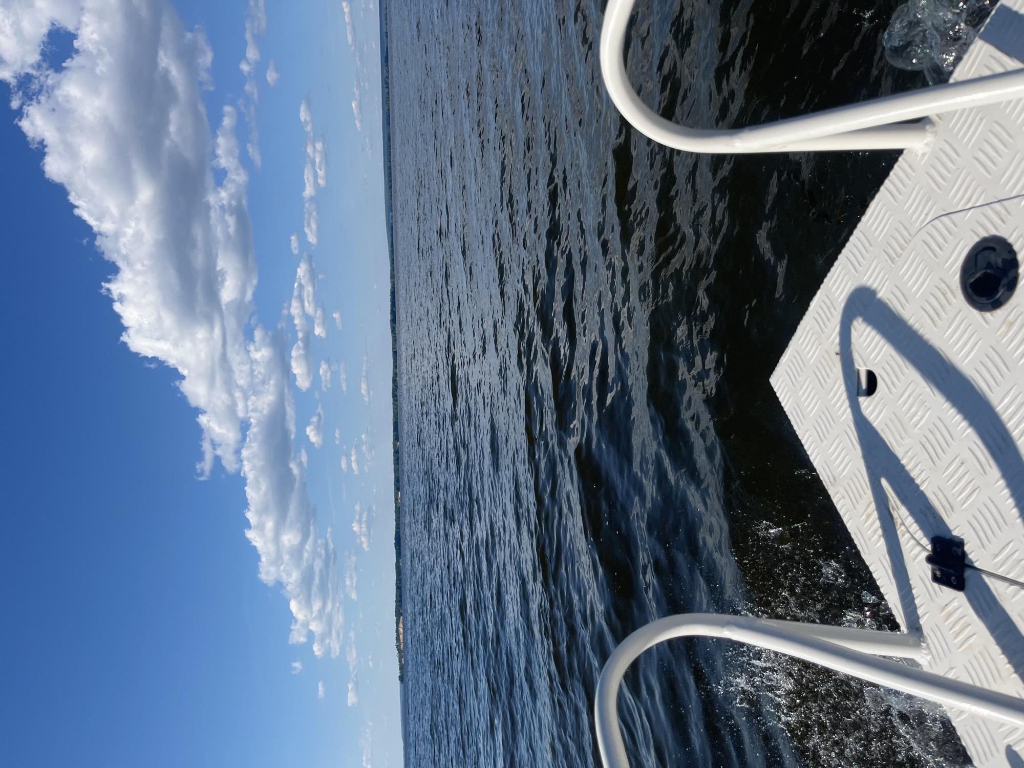 The image depicts a view from the bow of a white boat moving across a large body of dark water. The water is rippled, with white spray visible on the left side of the frame where the boat is cutting through the waves. In the foreground, the textured white deck of the boat is visible, along with white railings. The horizon is visible in the distance, showing a treeline and a small settlement on a gently sloping shore. Above the water, a bright blue sky is filled with scattered white cumulus clouds. The scene suggests a sunny day on the water, likely in a rural or natural setting. The location is identified as Siukterka, Russia.