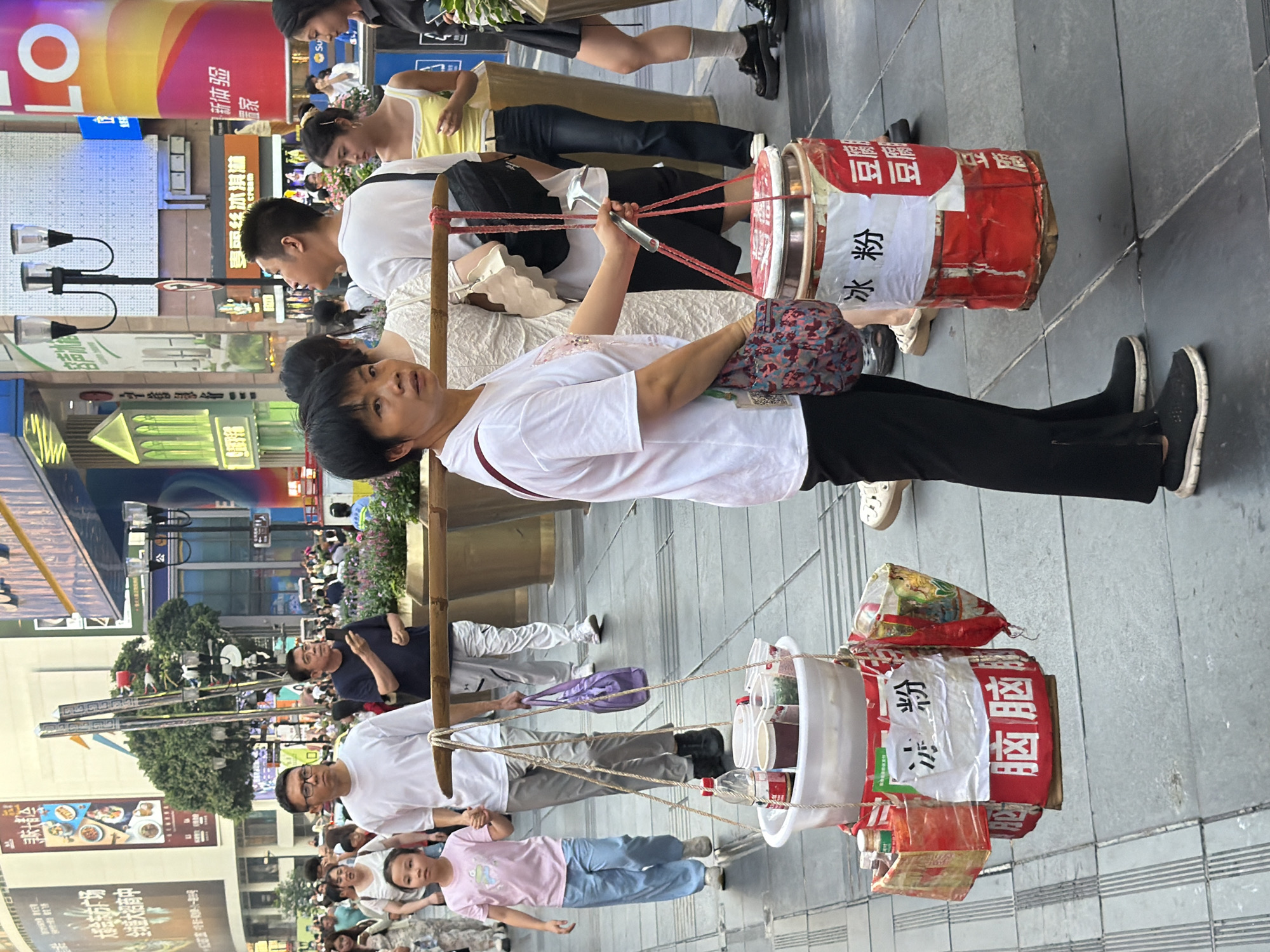 This image captures a street scene in Yuzhong District, China, with a woman carrying a yoke balanced on her shoulders. Suspended from the yoke are two large buckets containing food items. The woman is wearing a white t-shirt and black pants, with a patterned bag slung across her chest. She is walking on a paved sidewalk, surrounded by other pedestrians. In the background, buildings with various signs and advertisements are visible, suggesting a bustling urban environment. The overall atmosphere appears to be that of a busy commercial street.