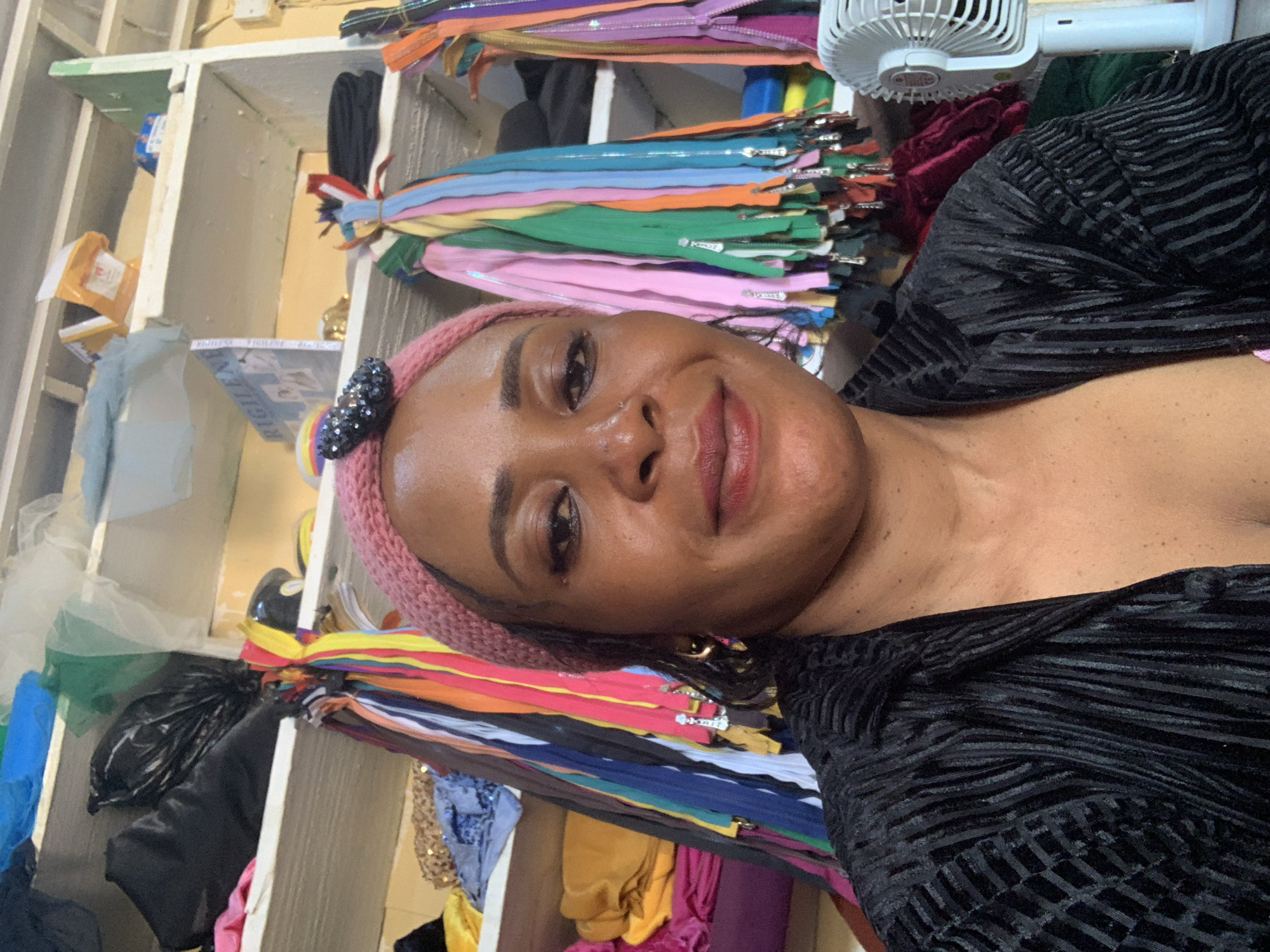 The image shows a close-up selfie of a woman in what appears to be a fabric or tailor shop in Yelwa, Nigeria. The woman is wearing a pink knitted hat with a black jeweled embellishment and a black textured shirt. Behind her are shelves filled with various fabrics and colorful zippers. The environment suggests a commercial space dedicated to sewing and fabric supply. The lighting is adequate, highlighting the woman's face and the surrounding materials.