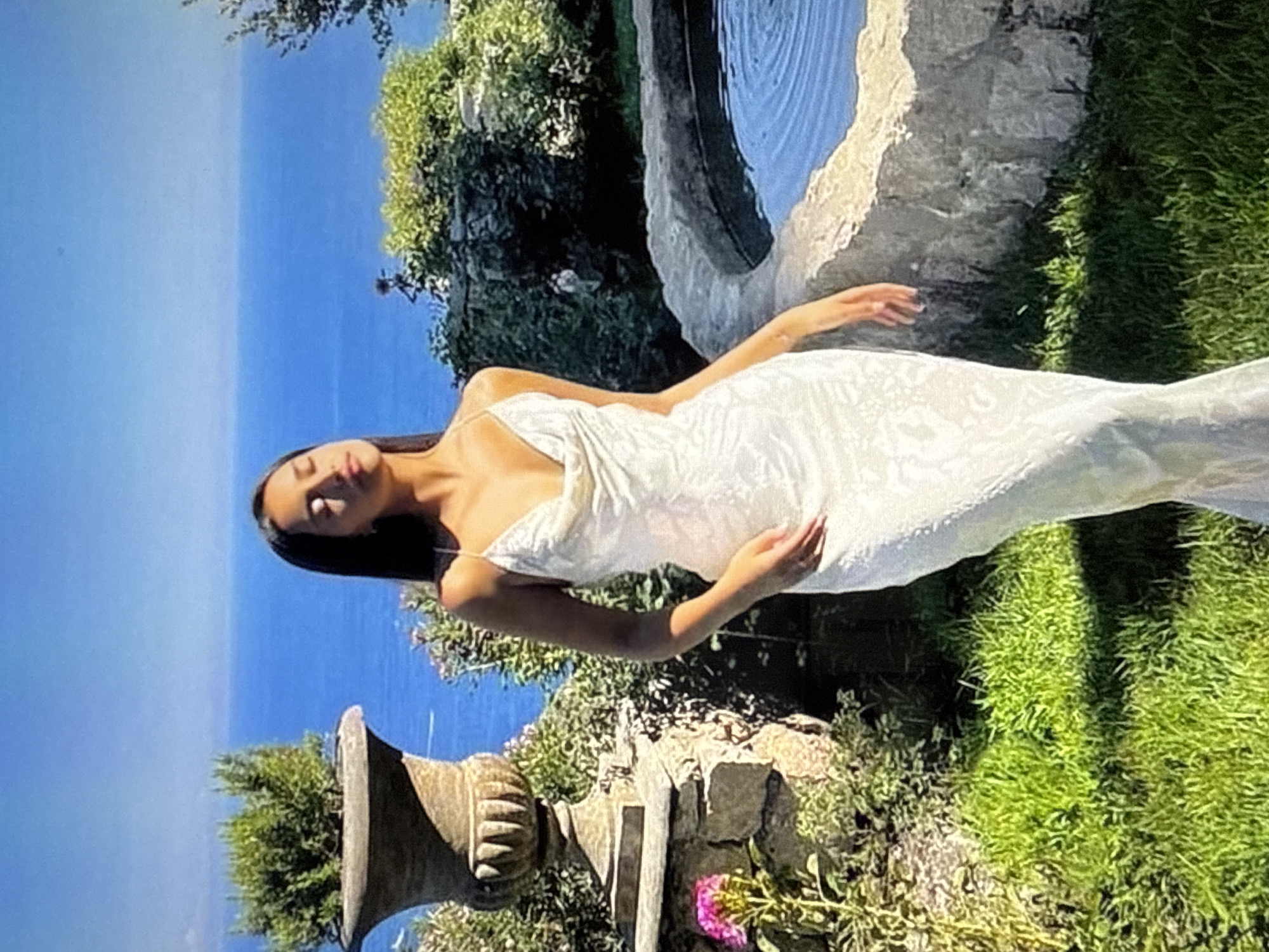 A young woman stands in a sun-drenched garden overlooking the sea. She is wearing a long, white, patterned dress with thin straps. Her eyes are closed, and her head is tilted back slightly, as if enjoying the sun. The garden is lush with green grass, flowering plants, and stone structures. In the background, the bright blue sea stretches to the horizon under a clear sky. The scene is peaceful and idyllic. This image is not located in Găvănoasa, Moldova.