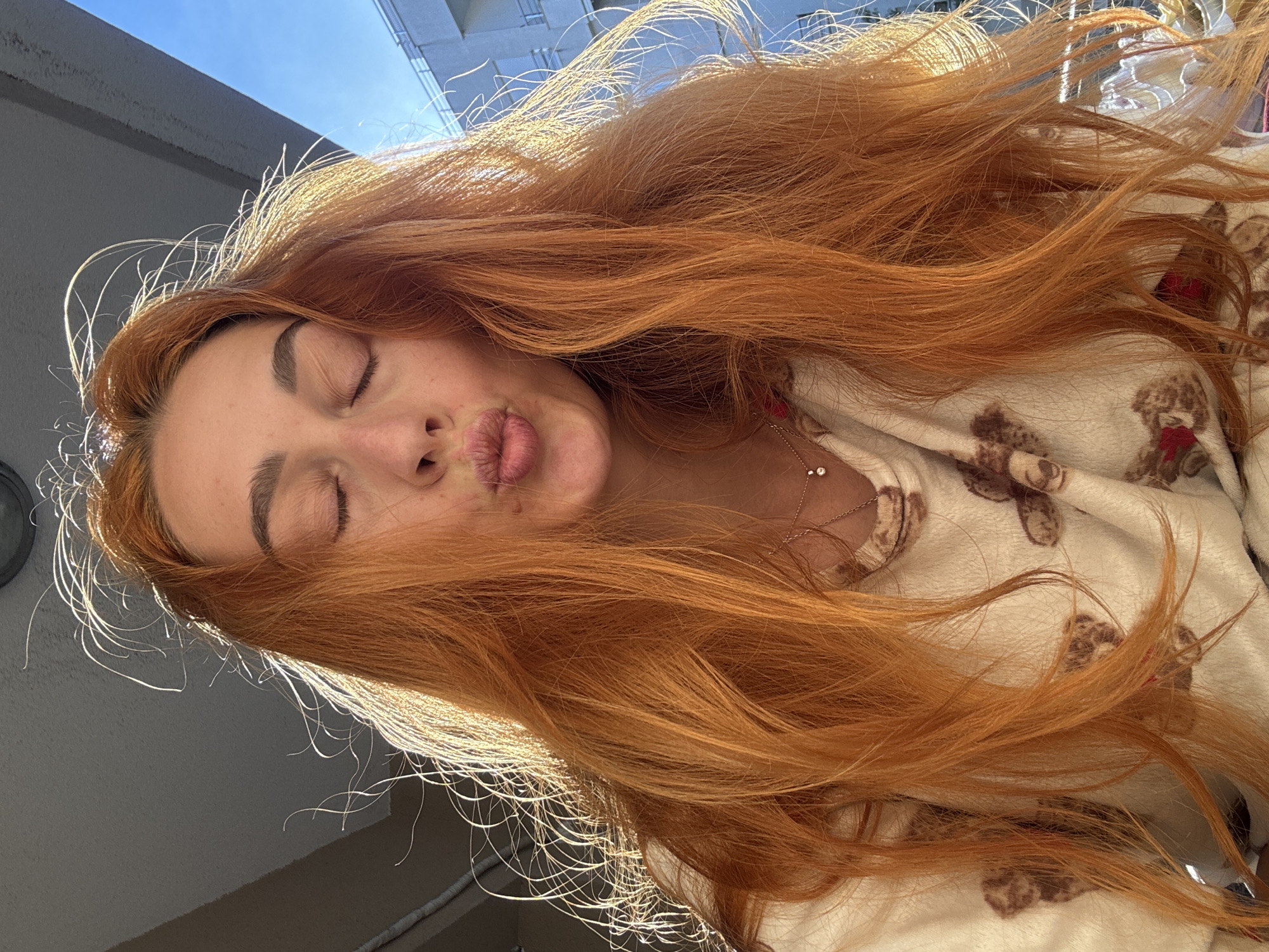 A young woman with closed eyes and pursed lips poses for a portrait, her auburn hair cascading across her face and shoulder. She wears a light-colored top with a teddy bear pattern. A delicate necklace adorns her neck. The background reveals a sunny sky, the top of a building, and a gray wall. The location appears to be Budva, Montenegro, as indicated by the architectural style and light typical of the region.