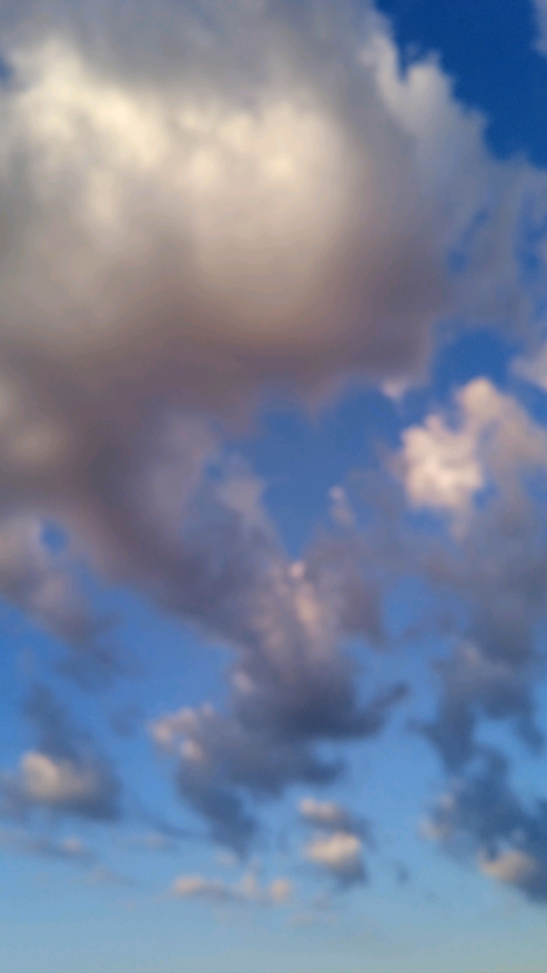The image depicts a sky filled with clouds against a blue background. The clouds are varied in size and color, with some appearing white and fluffy, while others are darker and more grey, suggesting depth and varying densities. The overall impression is of a dynamic sky, possibly before or after a weather event. The image is heavily blurred, giving it an abstract or impressionistic quality. There are no discernible landmarks or elements that would indicate a specific location within a city.