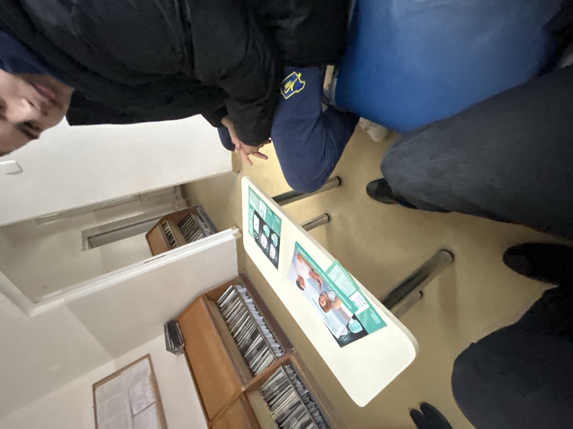 The image depicts an individual wearing a dark jacket and blue Nike sweatpants sitting on a blue chair. They are at a table with advertisements, potentially in Zlatograd, Bulgaria, based on the advertisements displayed. A cabinet filled with papers is nearby. Another person, wearing dark pants and shoes, sits adjacent. The location appears to be indoors, possibly an office or public waiting area. The photograph is taken from an unusual angle.