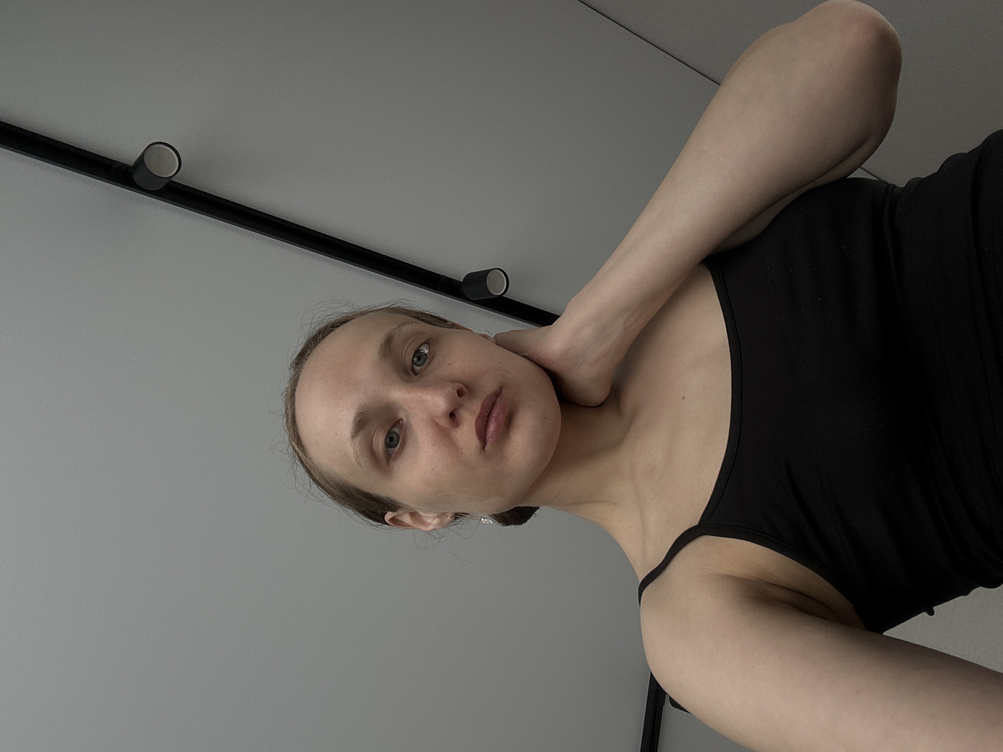 A medium-angle, close-up shot depicts a fair-skinned woman wearing a black tank top. She is oriented to the right of the frame, with her head tilted to the left and her hand resting on her neck. Her facial expression is neutral. The background consists of a gray wall with a black lighting track containing two cylindrical spotlights. The photo's origin or connection to Подшивалово, Russia, is not apparent from the visual information provided.