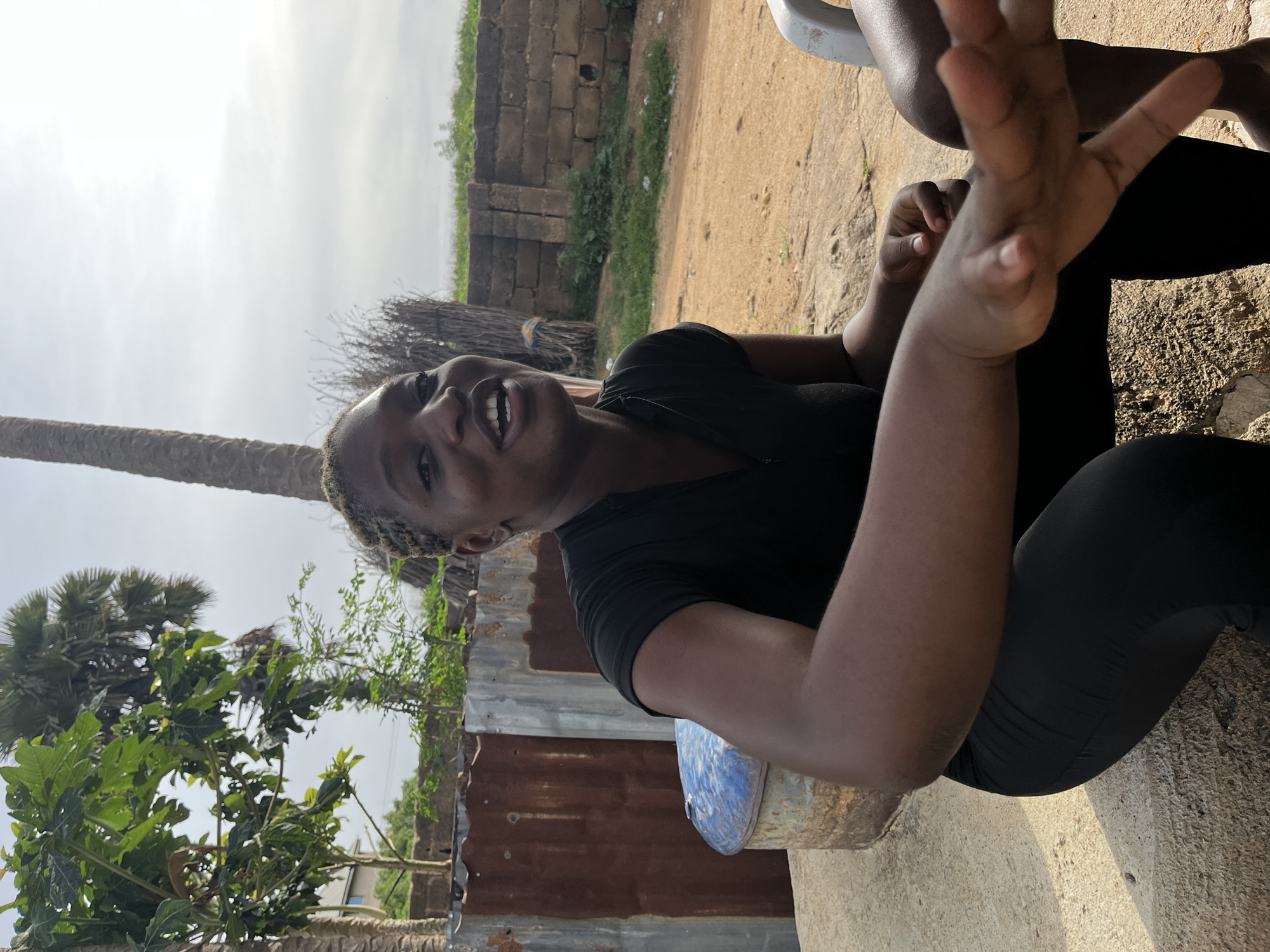 In an outdoor setting in Jos, Nigeria, a young Black woman with braided hair is seated outdoors, captured from the chest up. She is wearing a black t-shirt and black leggings, and is in the process of speaking, her mouth open and her expression animated. Her left arm is extended forward, with her hand visible in the foreground.
Behind her, a weathered blue barrel sits to her left, and corrugated metal sheeting forms a rustic fence on her left and behind her. Further back, a stone wall with some vegetation growing around it marks the boundary of the property. The ground is dusty and uneven, with patches of dry grass. The sky above is overcast, suggesting a cloudy day. To the far left of the frame, a palm tree adds a tropical element to the scene. The overall impression is one of a casual, informal moment captured in a semi-urban, possibly rural, environment within Jos.